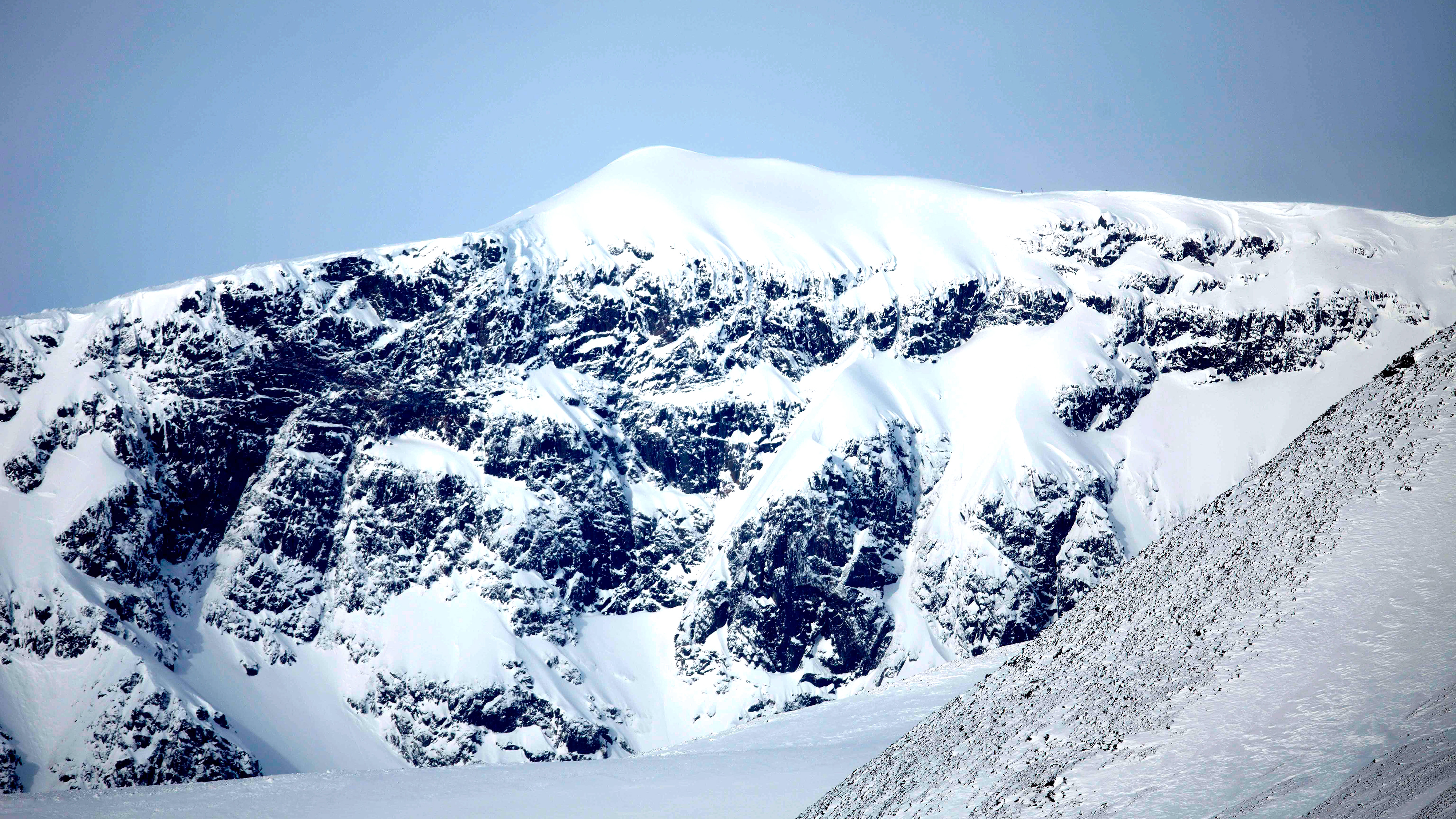 Snötäckt bergstopp med steniga partier under en blå himmel.