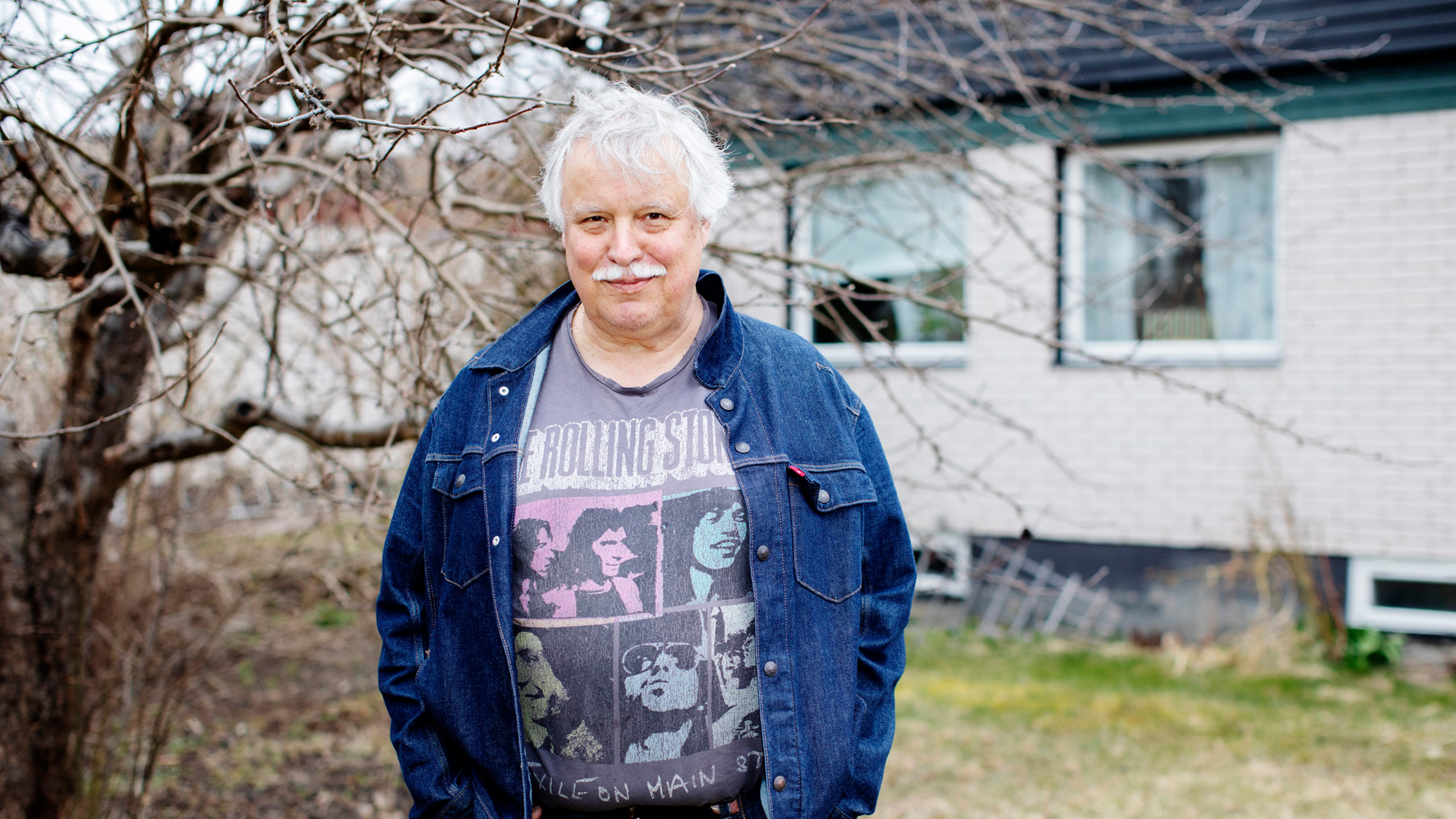 En äldre man med grått hår och mustasch står utomhus vid ett träd, har på sig en Rolling Stones T-shirt och jeansjacka. I bakgrunden syns ett hus och en gräsmatta.