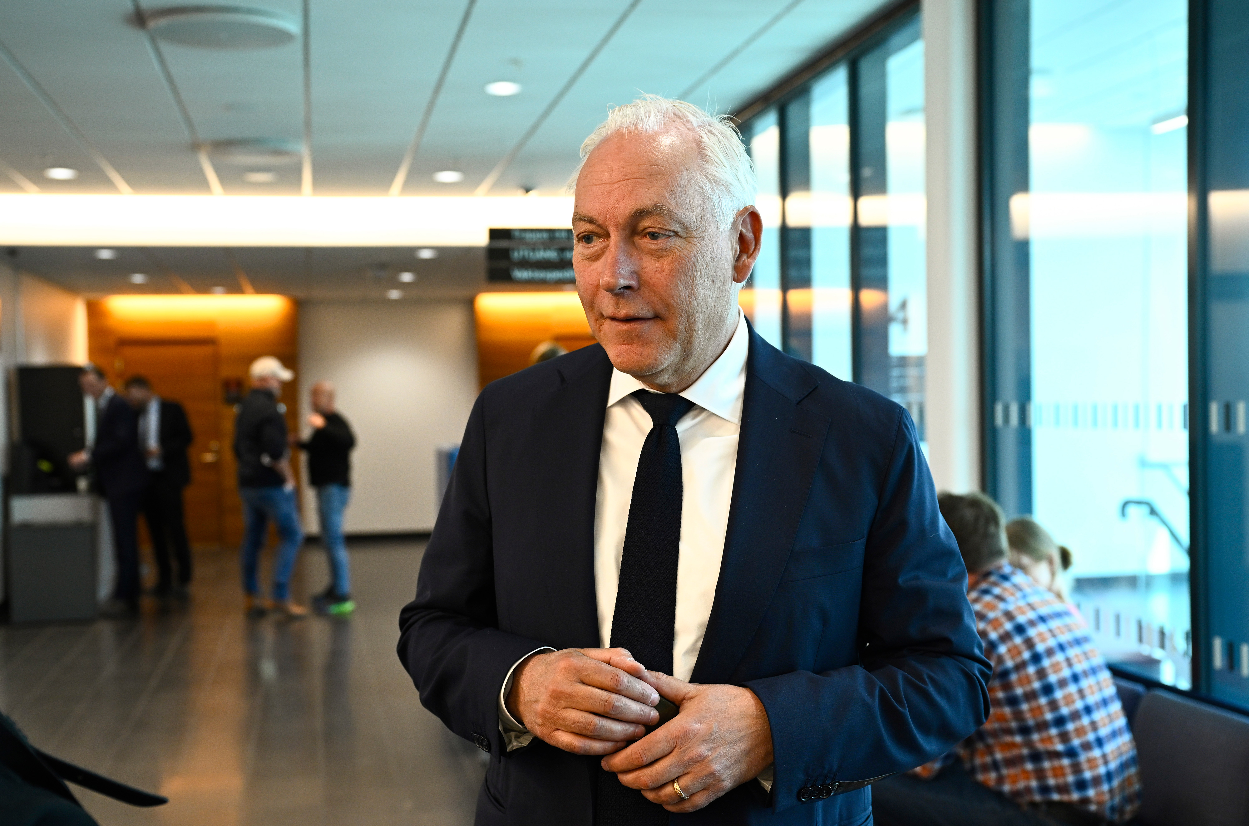 En man i kostym och slips står i en hall med glasväggar, medan andra personer syns i bakgrunden.