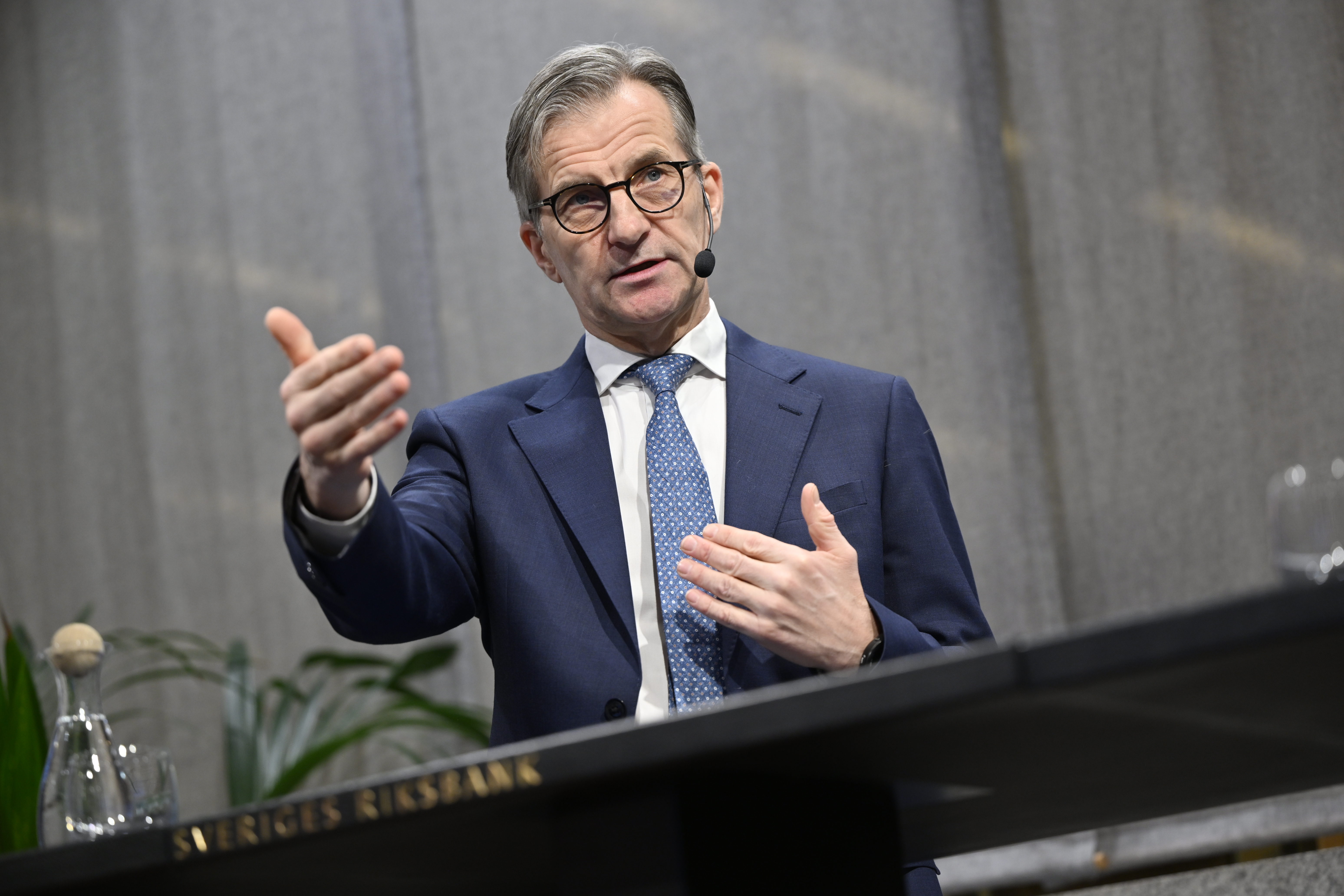 En man i kostym och slips talar i ett headset och gör en gest med ena handen vid ett podium märkt "SVERIGES RIKSBANK".