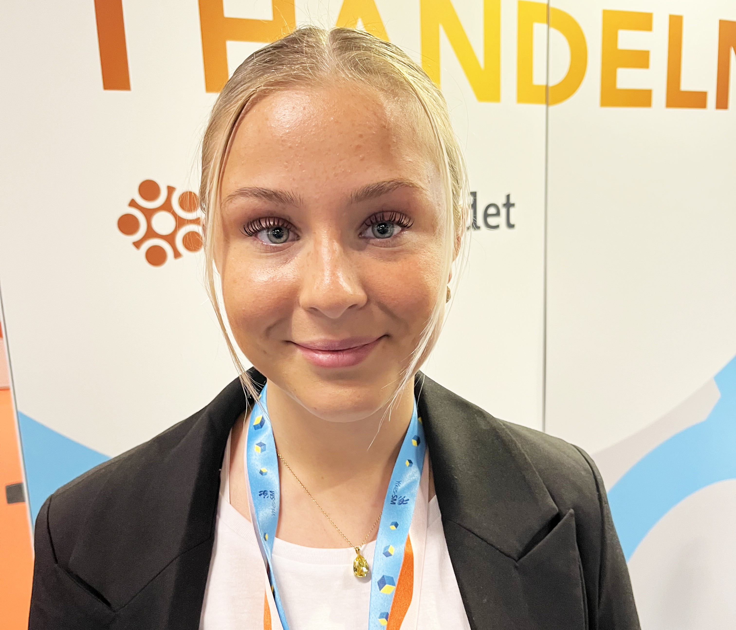 En kvinna med blont hår, svart kavaj och blått nyckelband står inomhus framför en skylt med orange text och en logotyp.