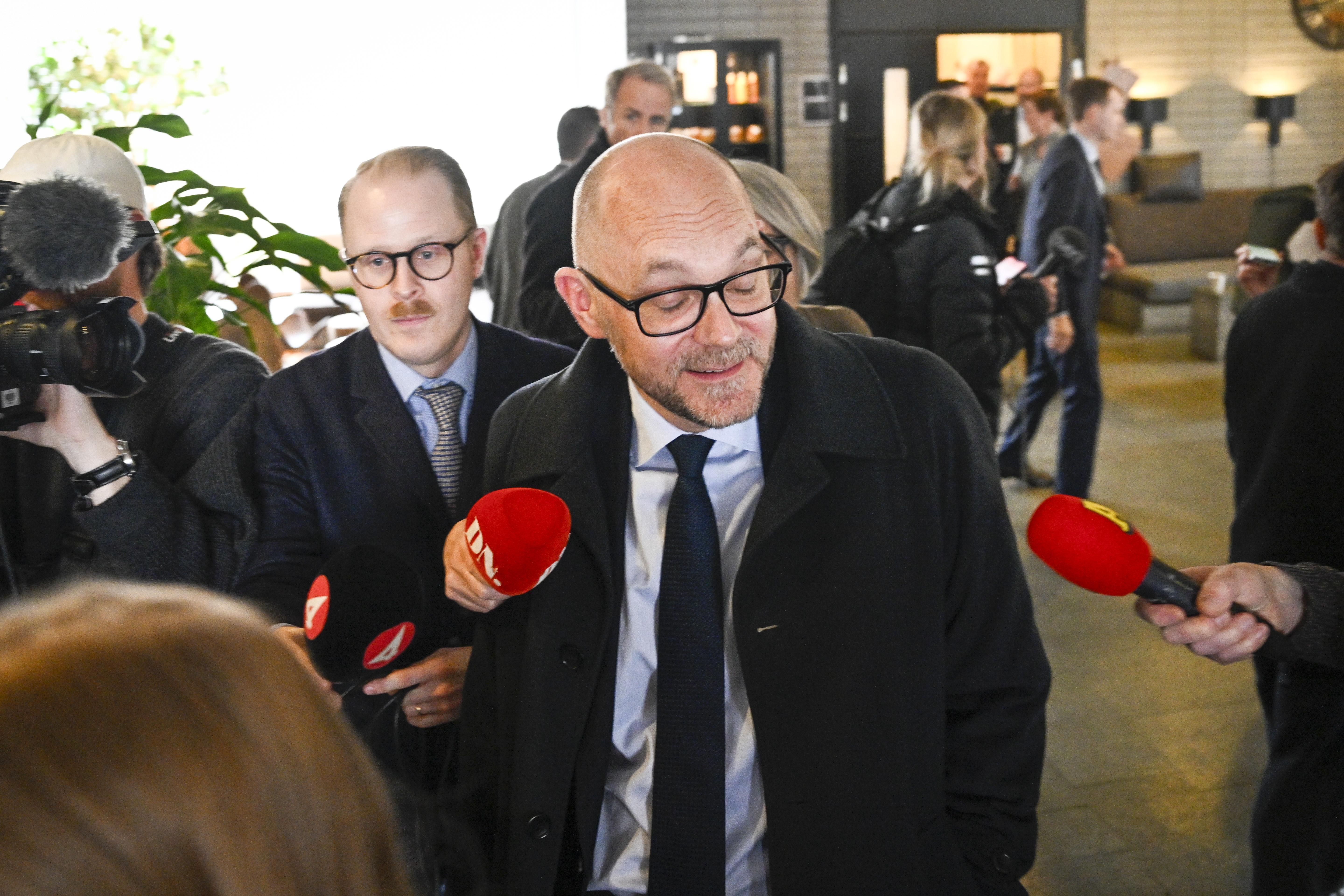 En man i kostym och glasögon omges av reportrar med mikrofoner med TV-logotyper. I bakgrunden står personer och pratar.
