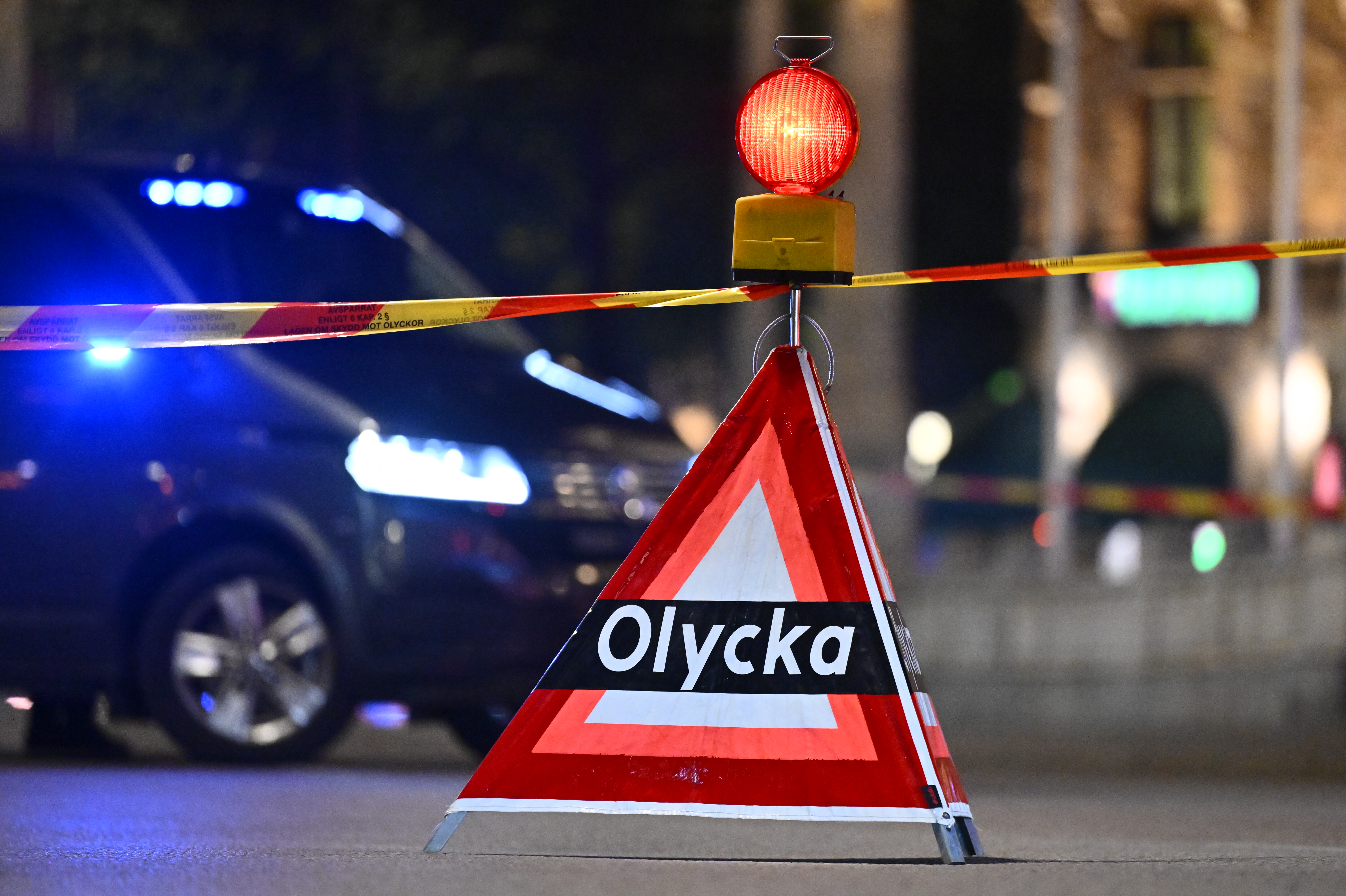 En varningstriangel med texten "Olycka" står framför polisavspärrning och ett fordon med blinkande ljus på natten.