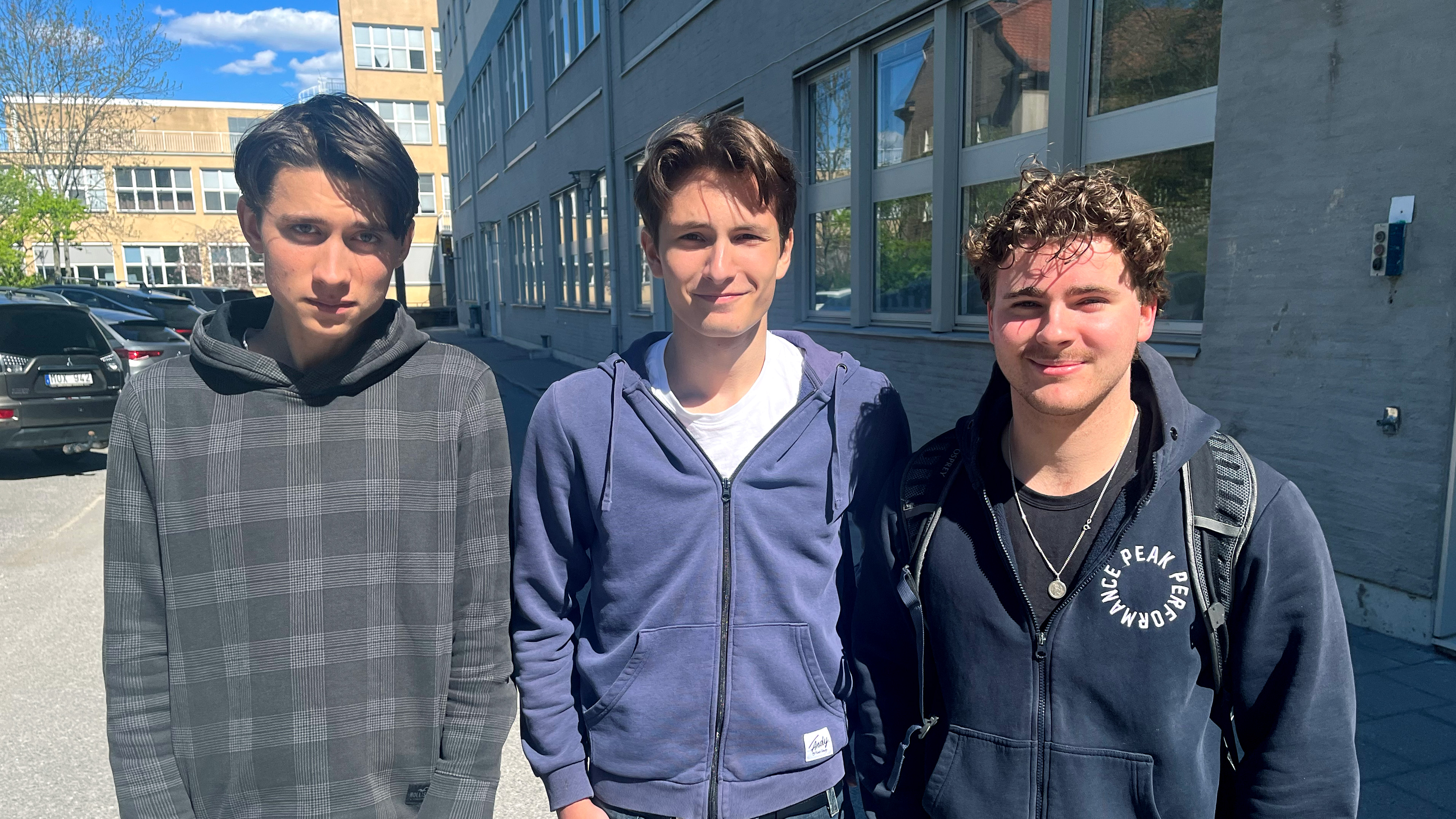 Tre unga män står utanför en byggnad. Alla har på sig huvtröjor eller jackor med huva.