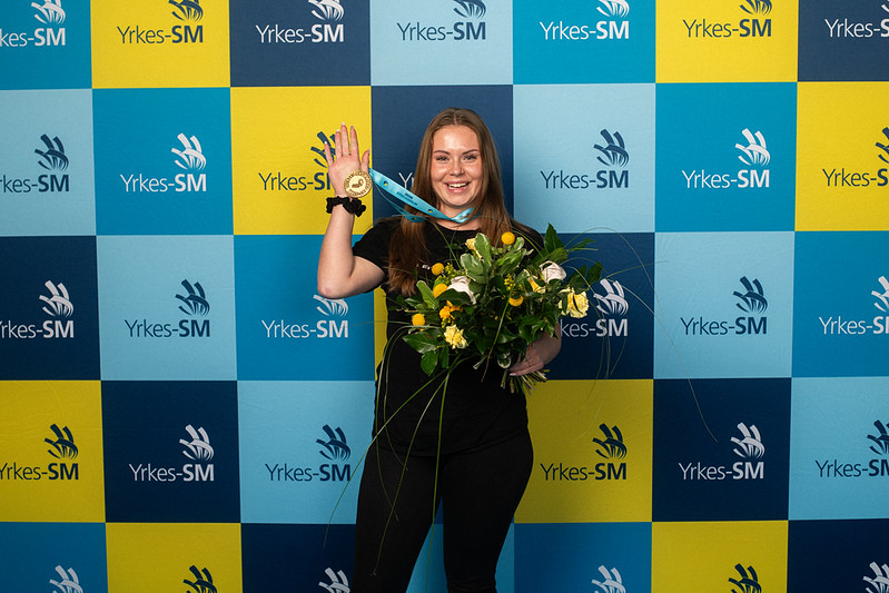 En kvinna står framför en professionell mästerskapsbakgrund, håller i en bukett blommor och visar upp sin medalj med ett band runt halsen.