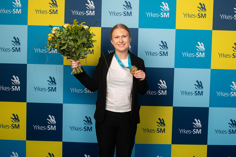 En person står framför en blå och gul professionell mästerskapsbakgrund, håller i en bukett blommor och en medalj och ler mot kameran.