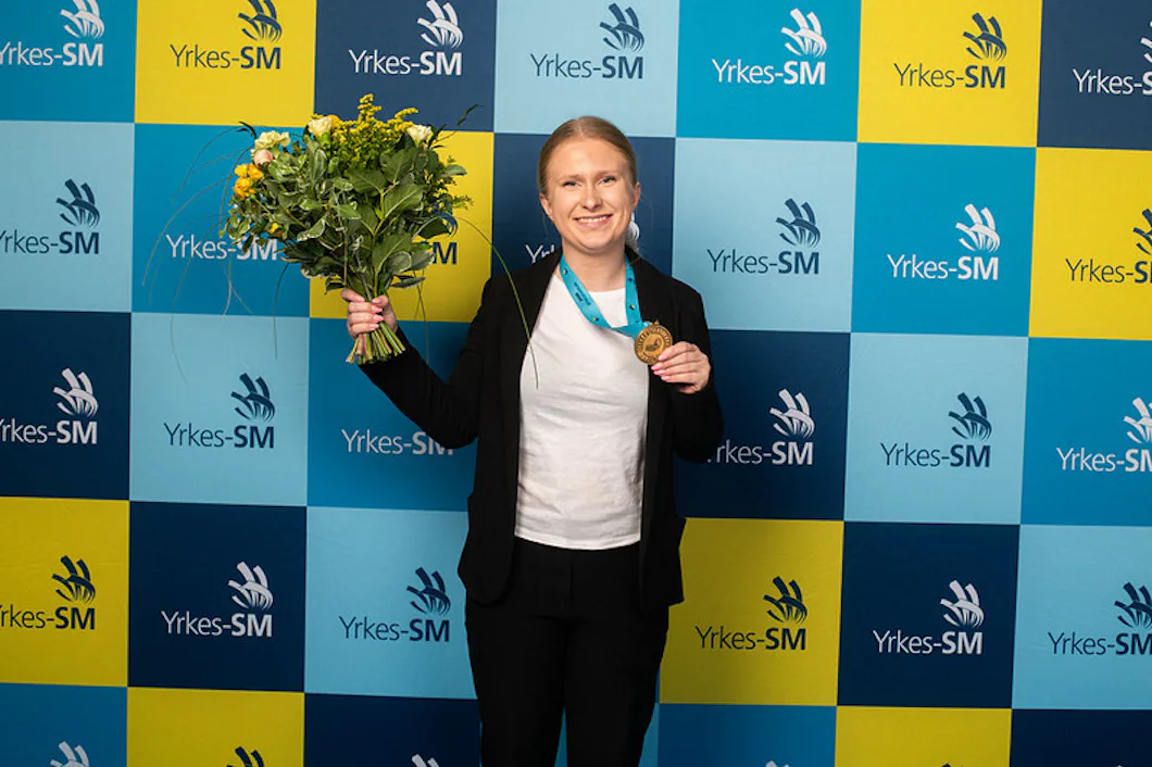 En person står framför en blå och gul professionell mästerskapsbakgrund, håller i en bukett blommor och en medalj och ler mot kameran.