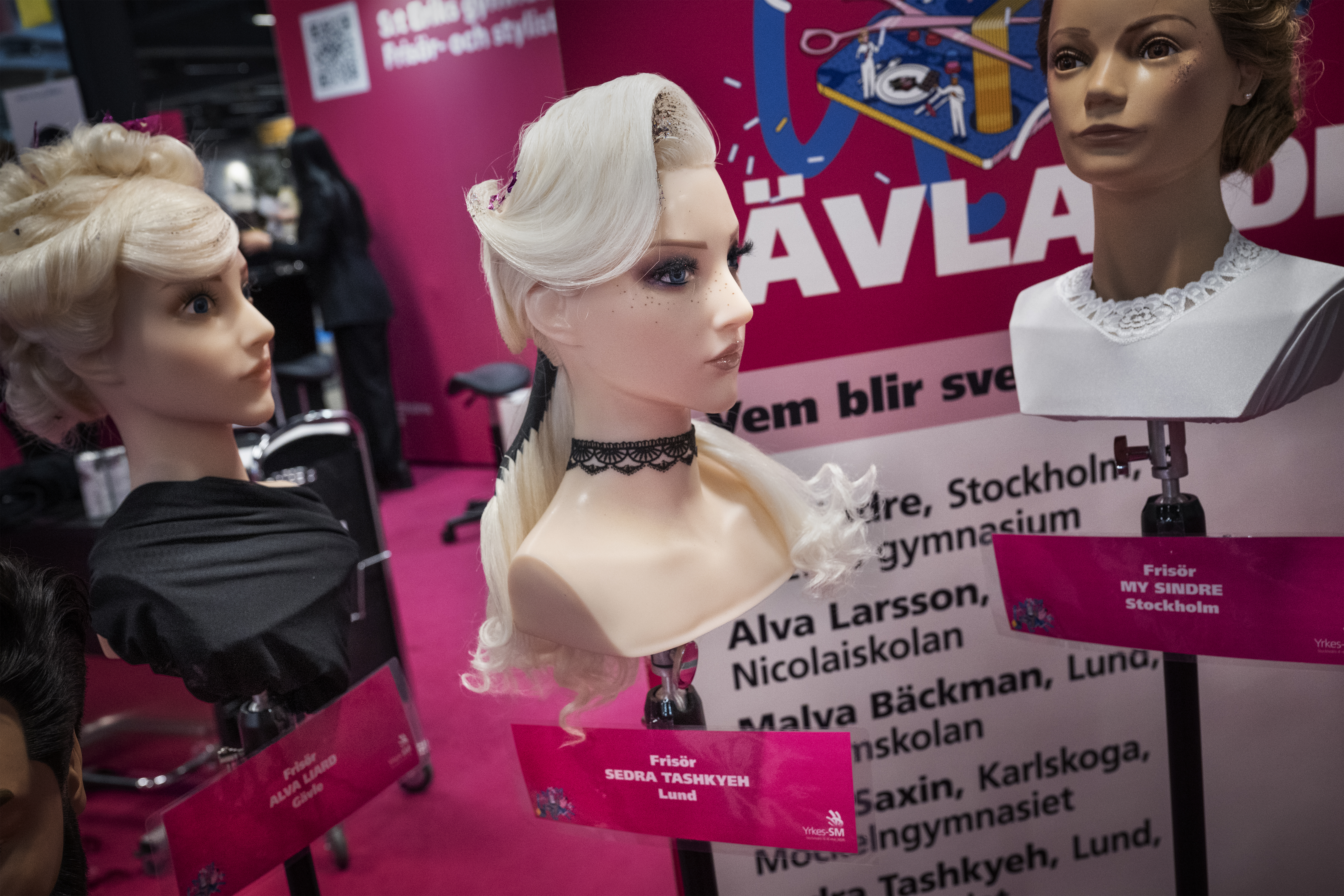 Tre skyltdockahuvuden med hår och smink är placerade på ett stativ framför en rosa bakgrund med svenska bokstäver och namnlappar.