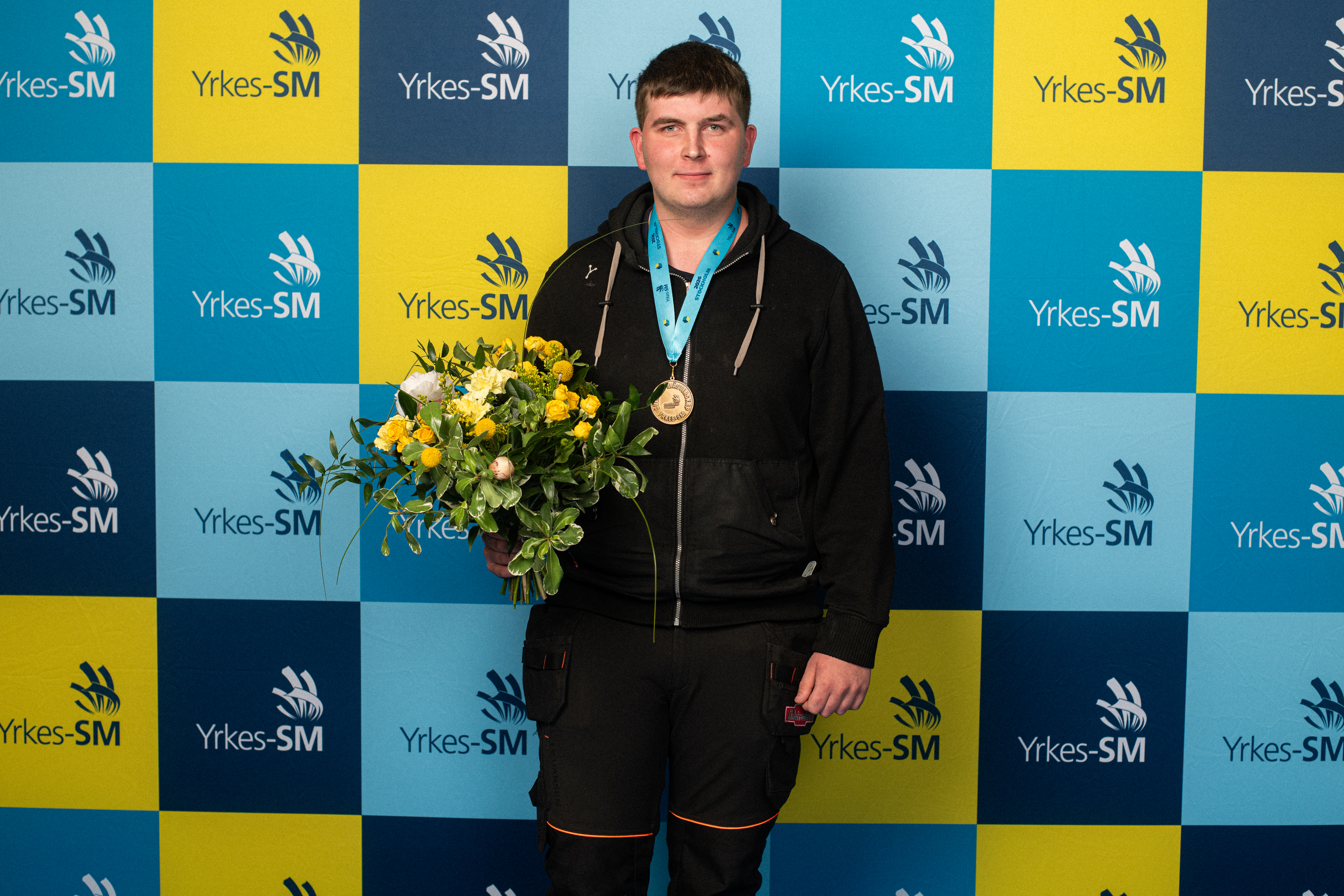 En man med svart jacka och medalj håller en bukett blommor framför en blå och gul Yrkes-SM-bakgrund.