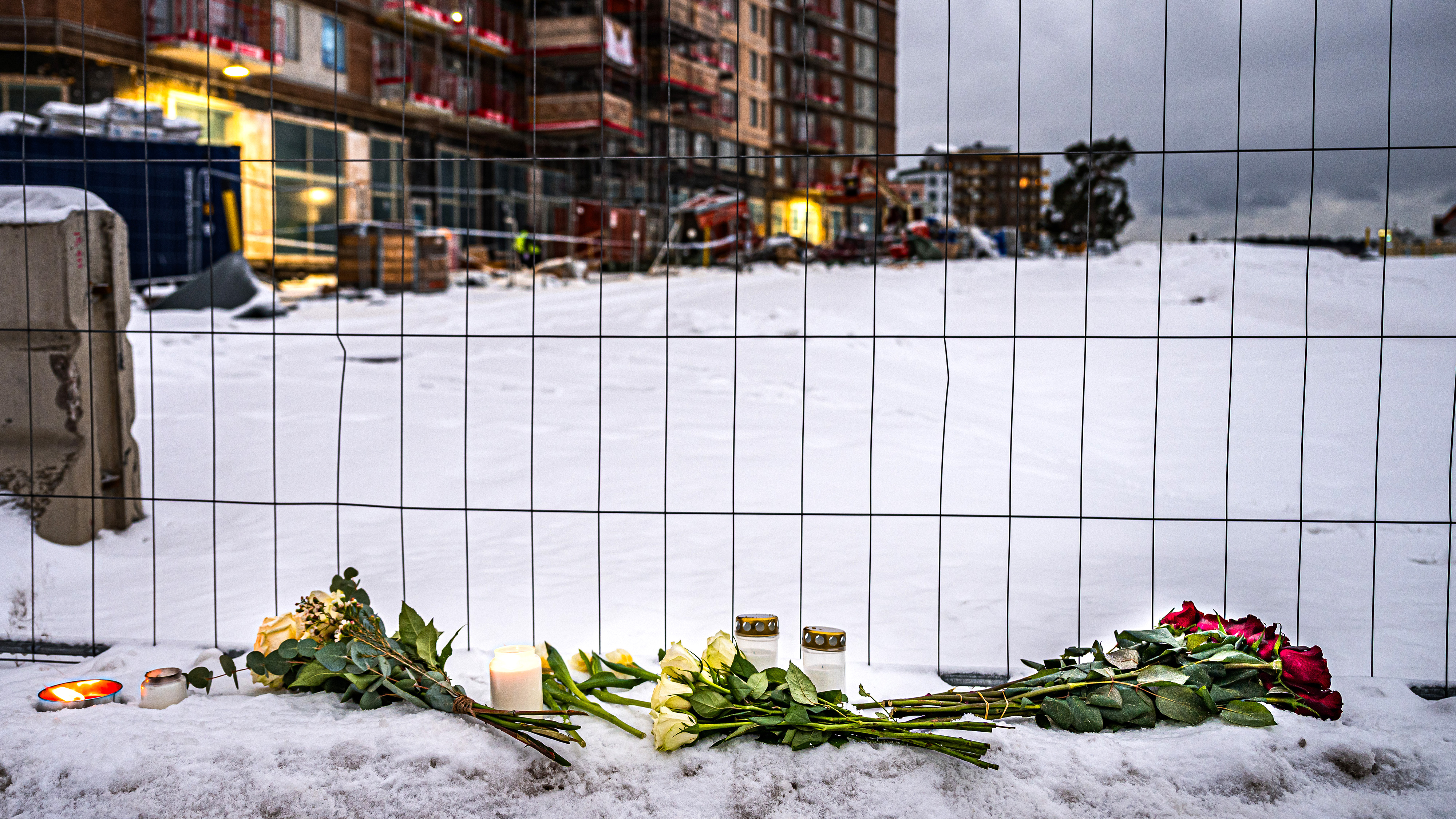 Blommor och ljus står i snön framför en byggarbetsplats bakom ett stängsel av metall.
