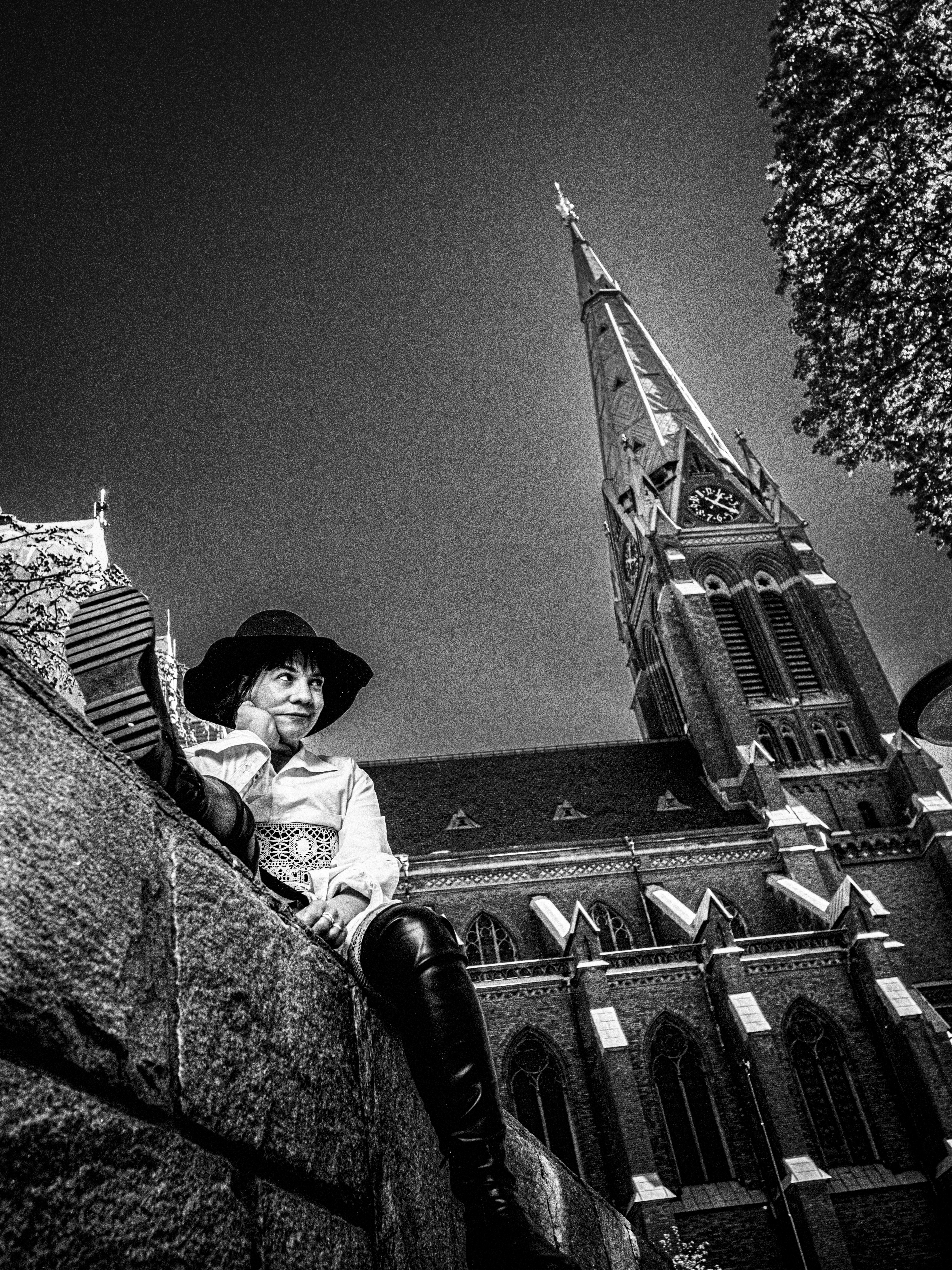 En kvinna i hatt och stövlar sitter på en stenmur framför en gotisk kyrka med ett klocktorn. Den är tagen i svartvitt.