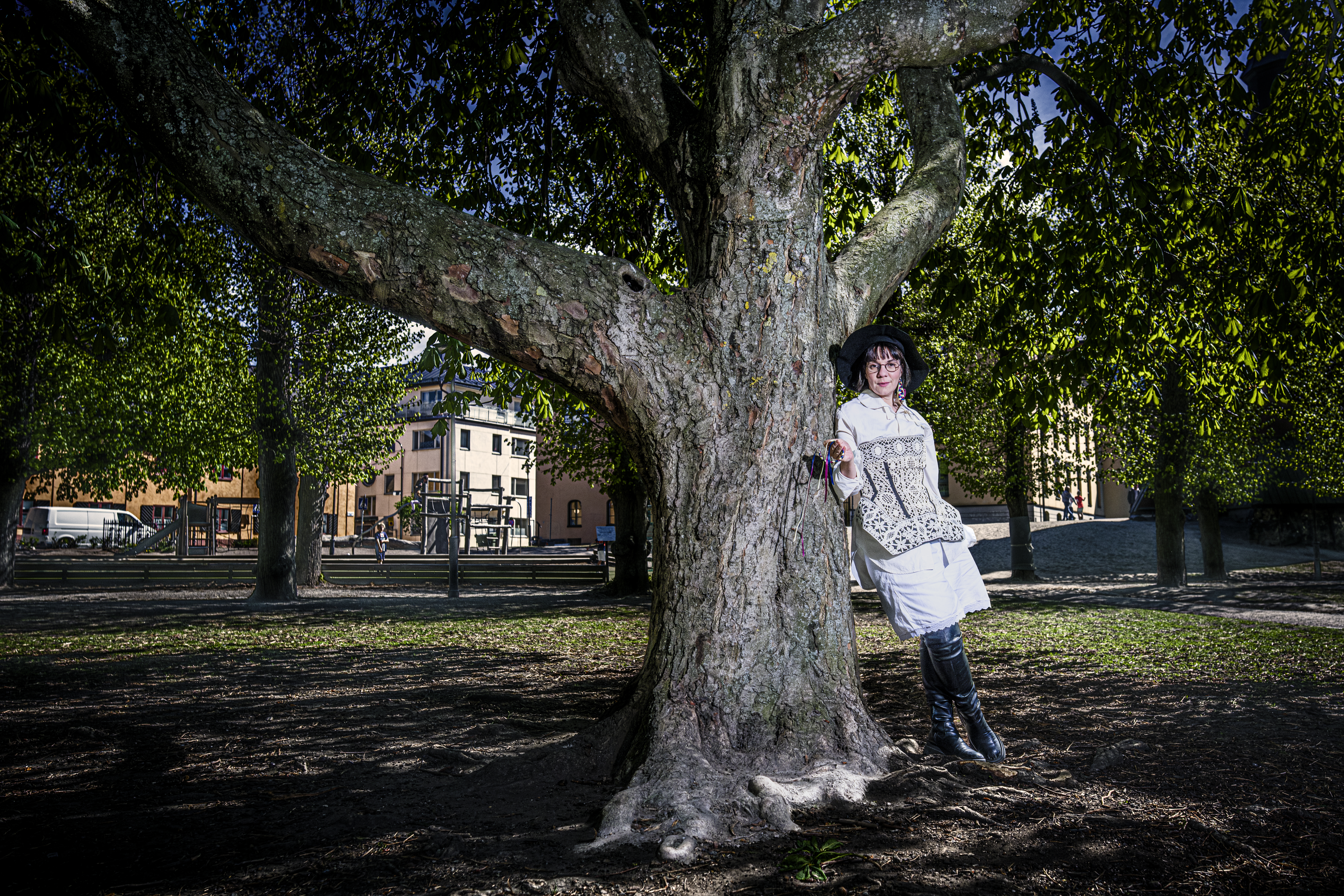 En person i vita kläder och en svart hatt står lutad mot ett stort träd i en park med byggnader synliga i bakgrunden.
