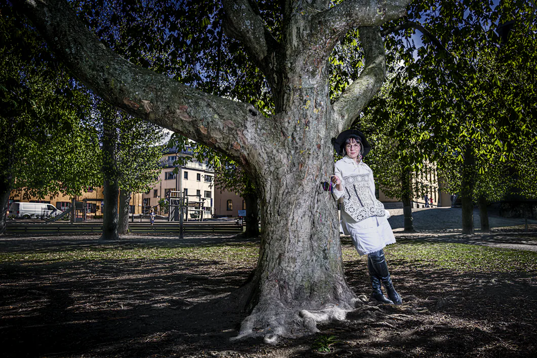 En person i vita kläder och en svart hatt står lutad mot ett stort träd i en park med byggnader synliga i bakgrunden.