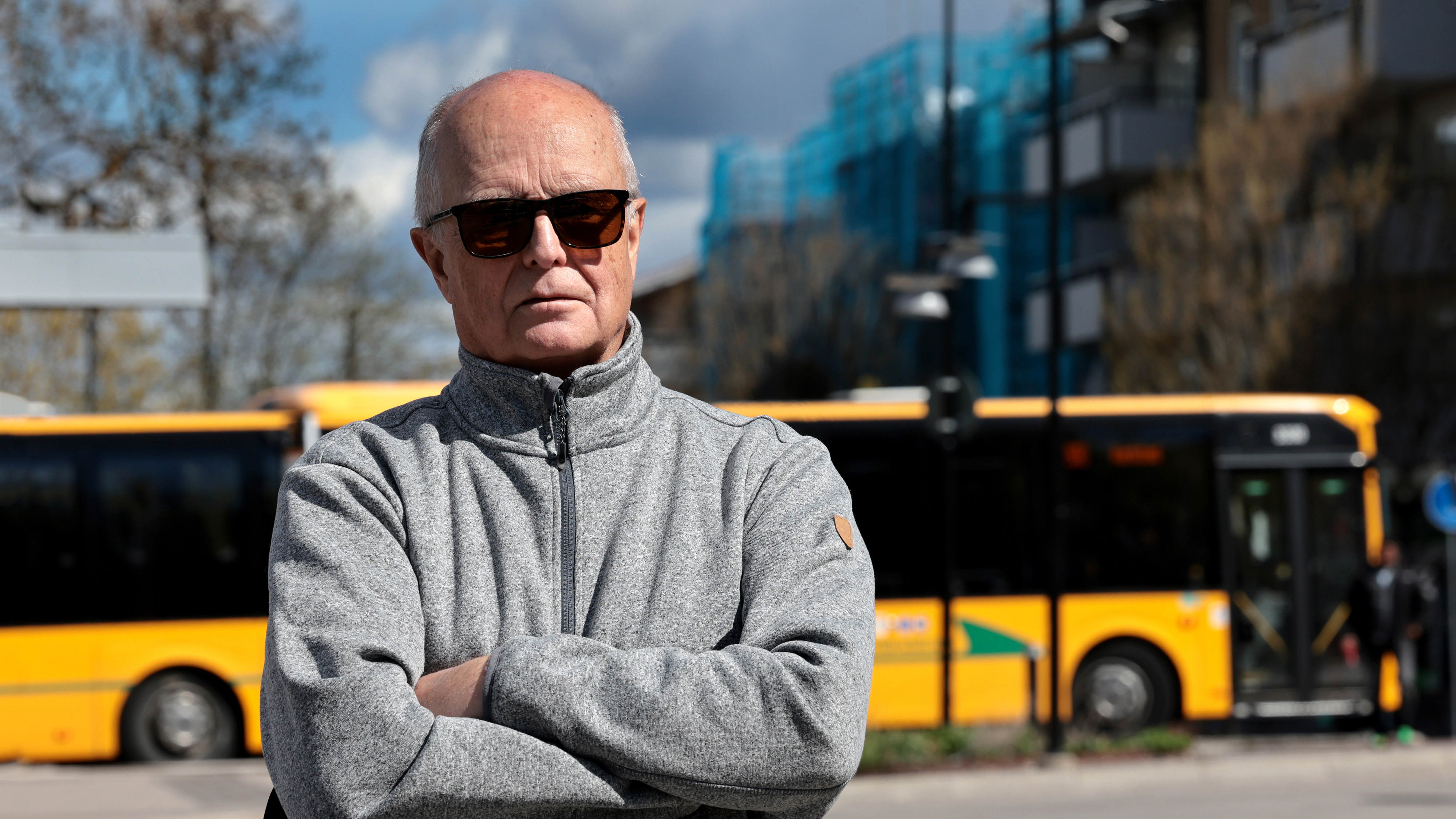 En äldre man med solglasögon och grå jacka står utomhus med armarna i kors. Gula stadsbussar och byggnader syns bakom.