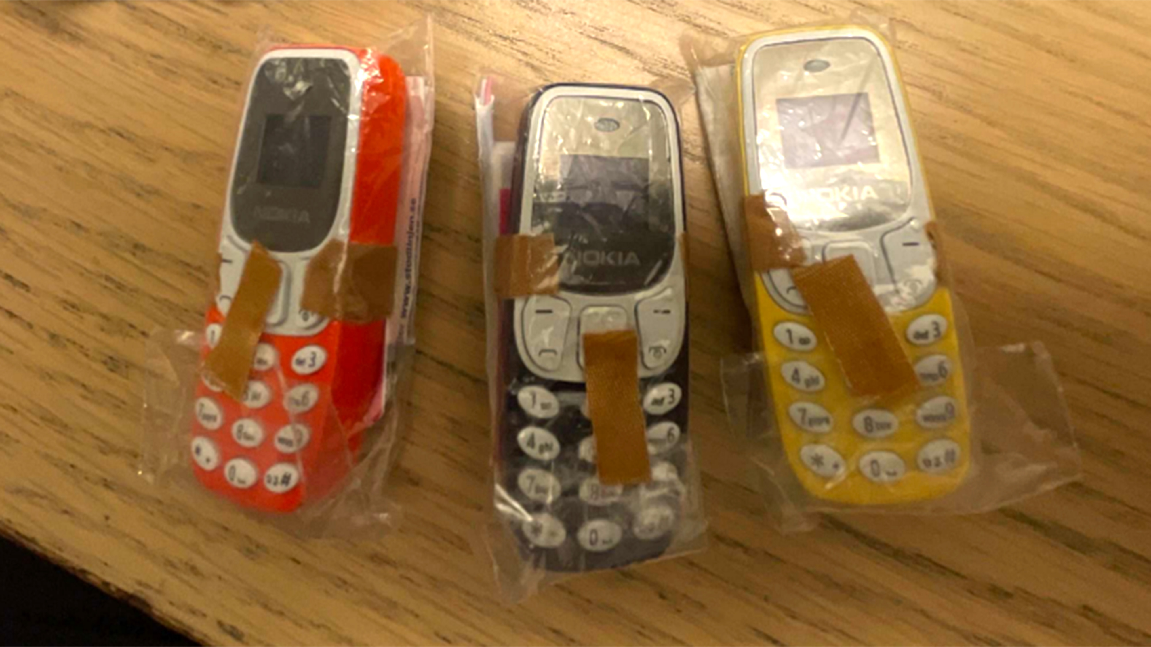 Tre små Nokia-telefoner inslagna i plast och tejp ligger bredvid varandra på en träyta.