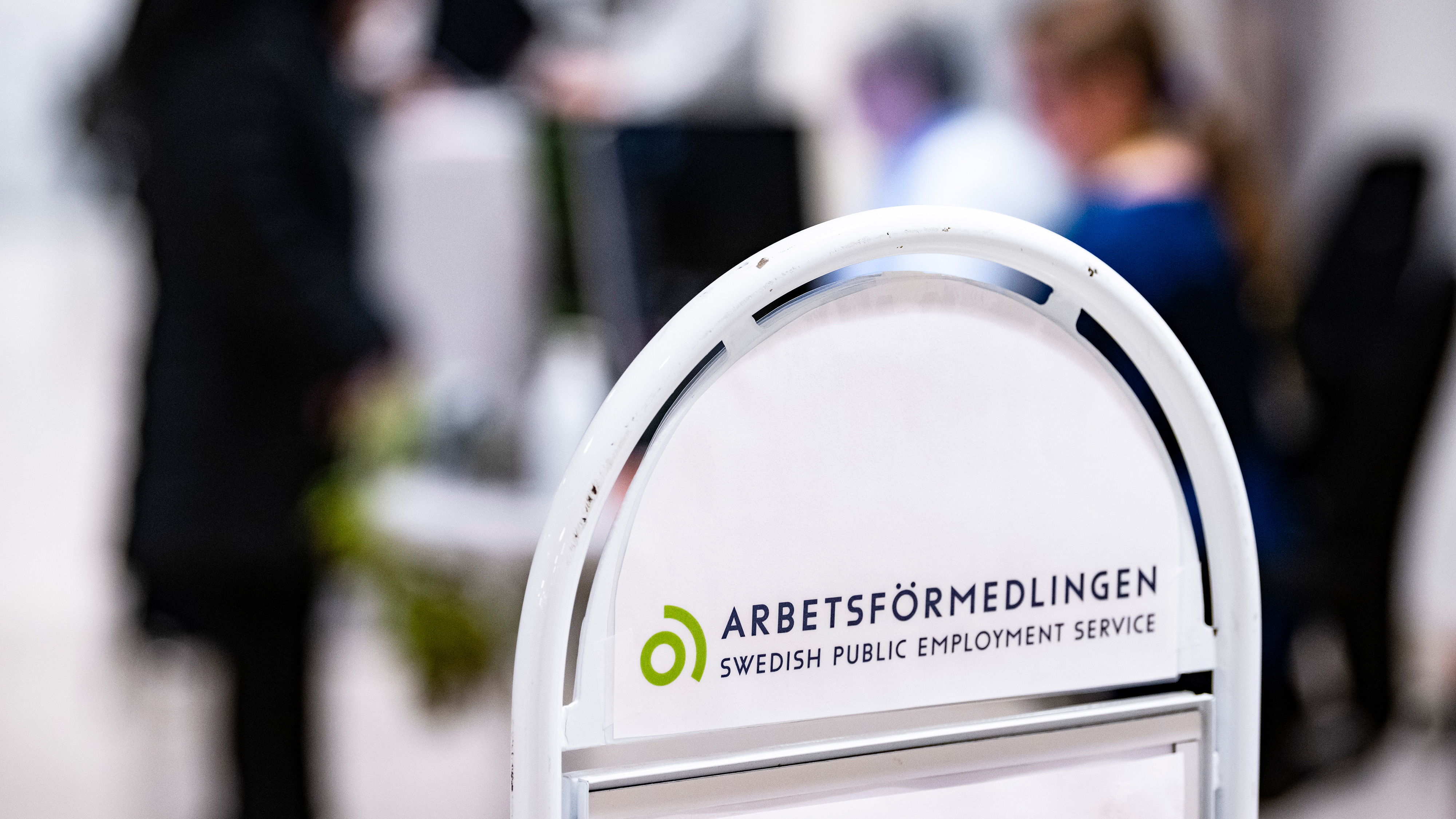 En skylt för Arbetsförmedlingen, den svenska arbetsförmedlingen, med personer i bakgrunden på ett kontor.