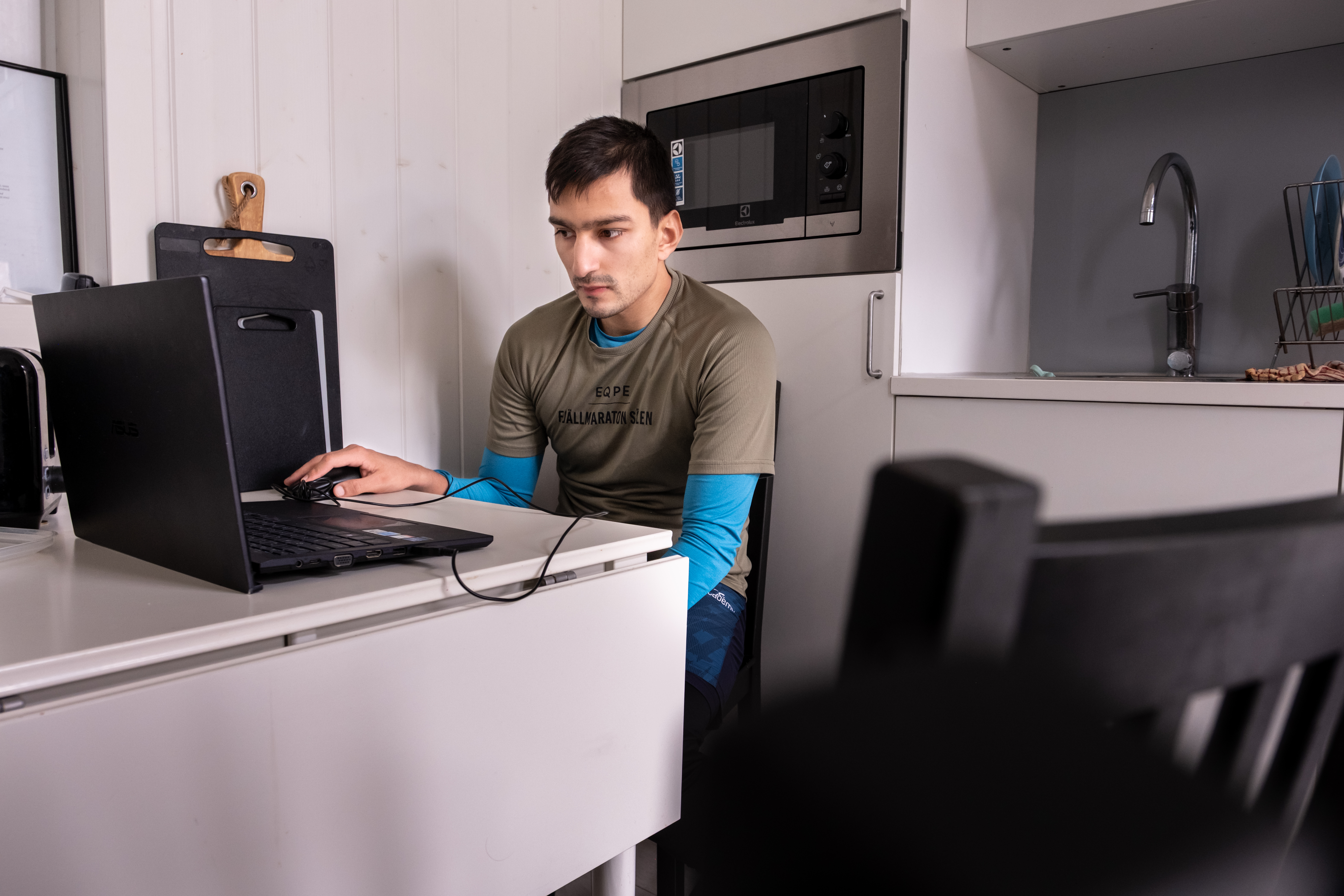 Oskar sitter vid en laptop som står på ett vitt borf. I bakgrunden syns en microvågsugn och en vattenkran.
