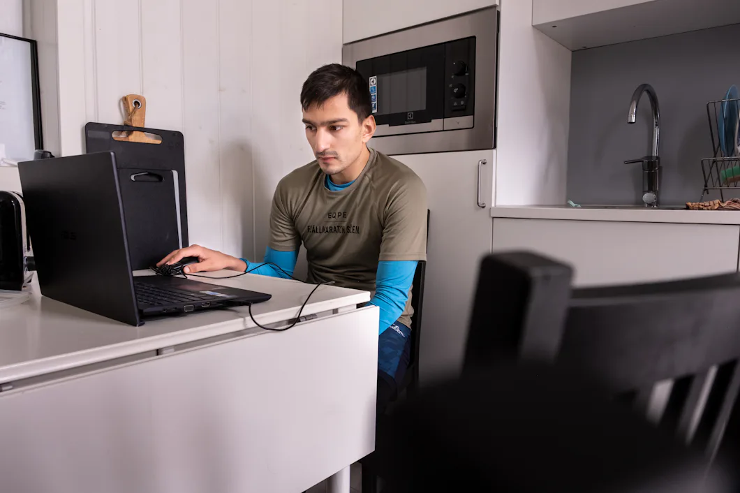Oskar sitter vid en laptop som står på ett vitt borf. I bakgrunden syns en microvågsugn och en vattenkran.