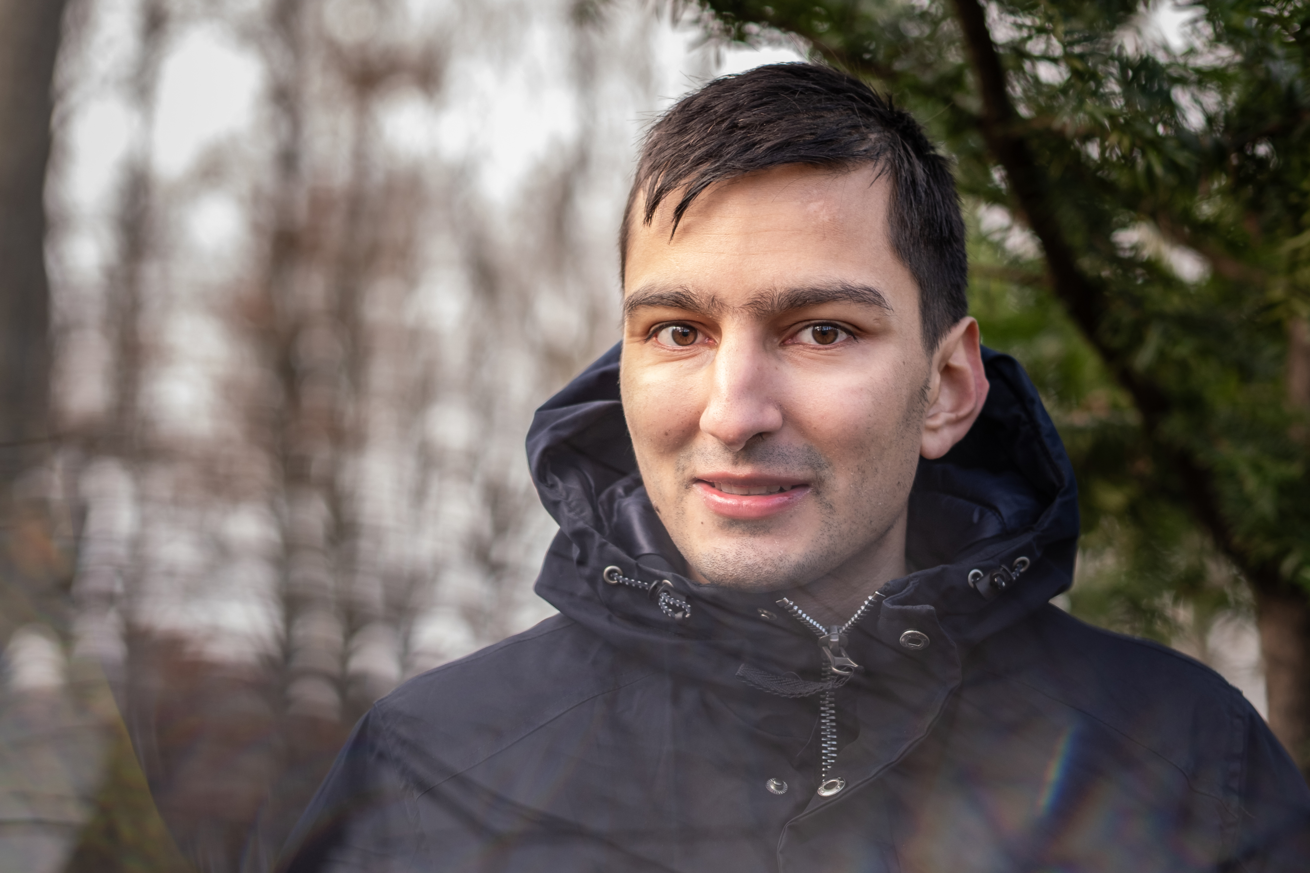 Oskar Bäckström med mörk jacka och huva står utomhus framför träd och tittar in i kameran med neutral min.