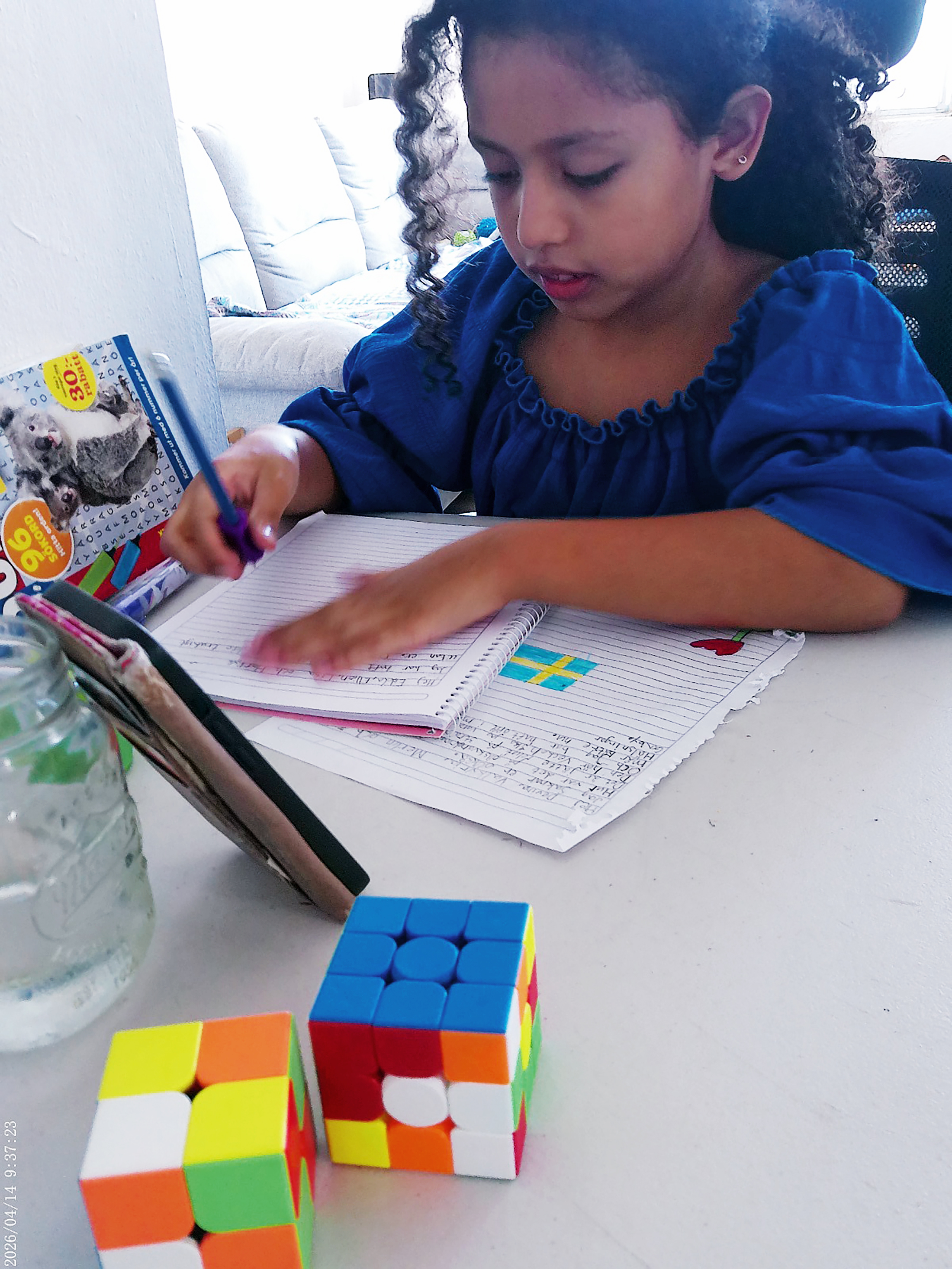 Gabby, iklädd en blå klänning, skriver i en anteckningsbok vid ett bord med två Rubiks kuber, en glasflaska och en öppen bok.