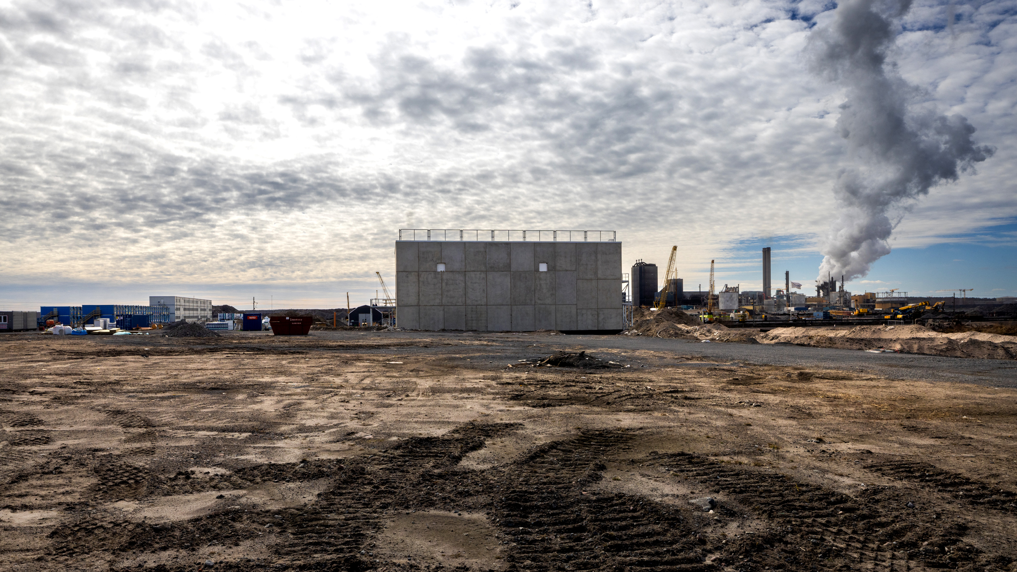 En byggarbetsplats med en betongstruktur i mitten, industribyggnader och rök i bakgrunden under en molnig himmel.