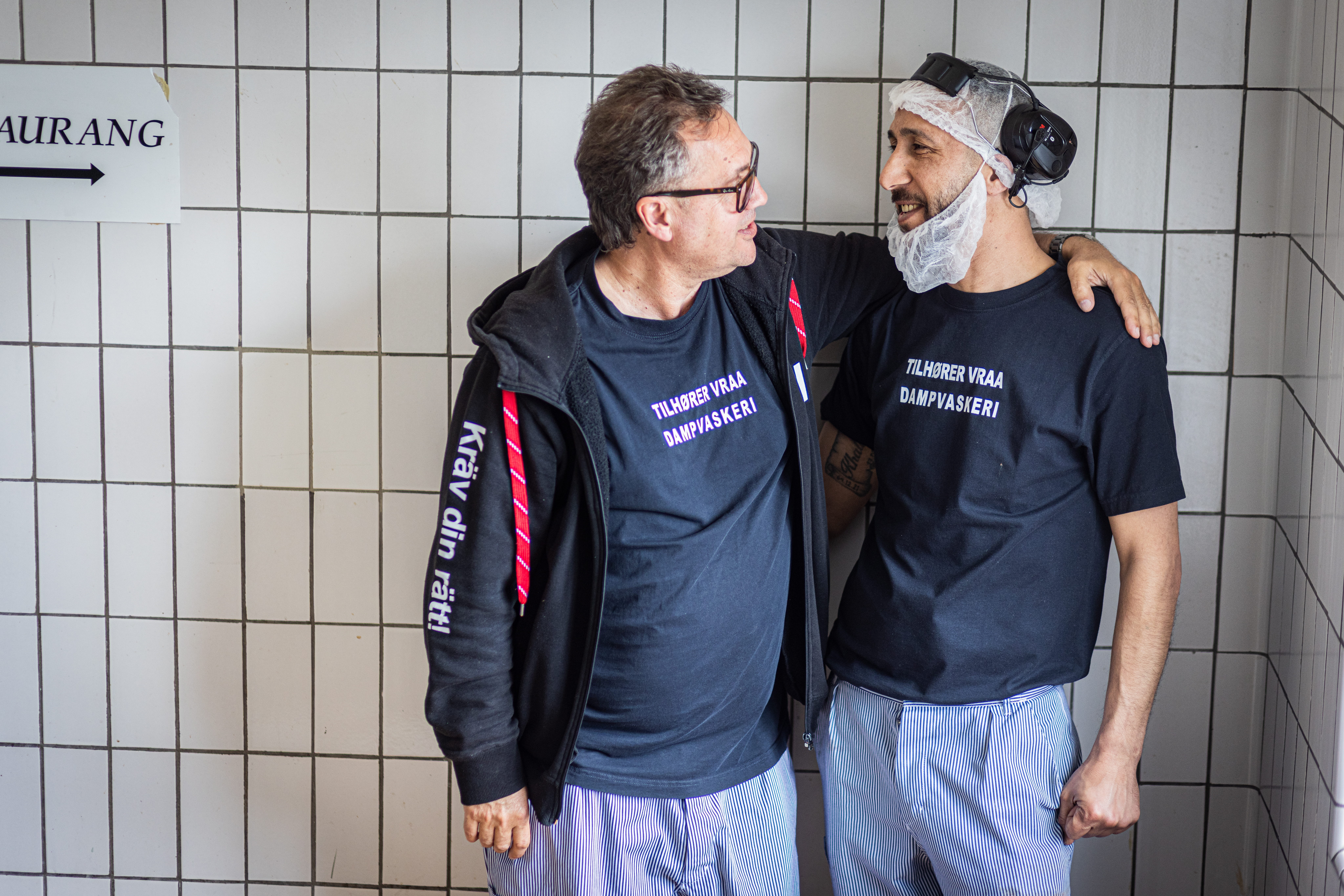 Två män klädda i svarta T-shirts, randiga byxor och hårnät står bredvid varandra i ett kaklat rum och pratar med armarna sammanlänkade.