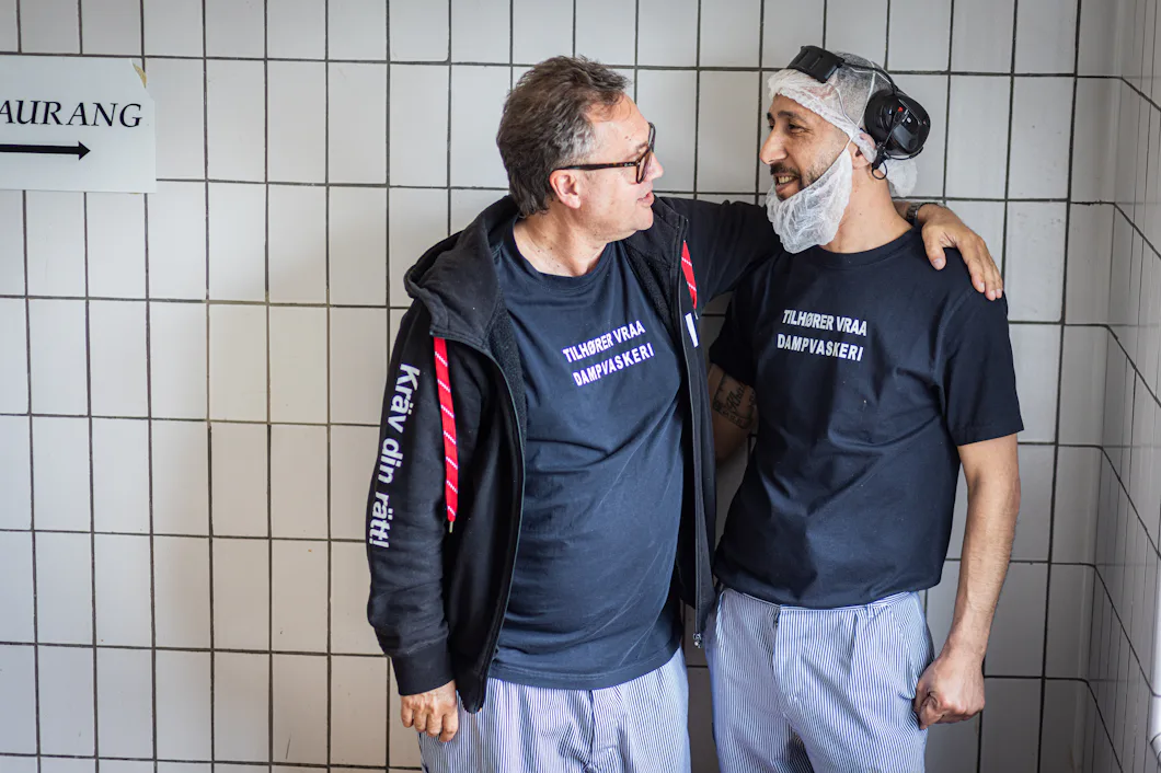 Två män klädda i svarta T-shirts, randiga byxor och hårnät står bredvid varandra i ett kaklat rum och pratar med armarna sammanlänkade.