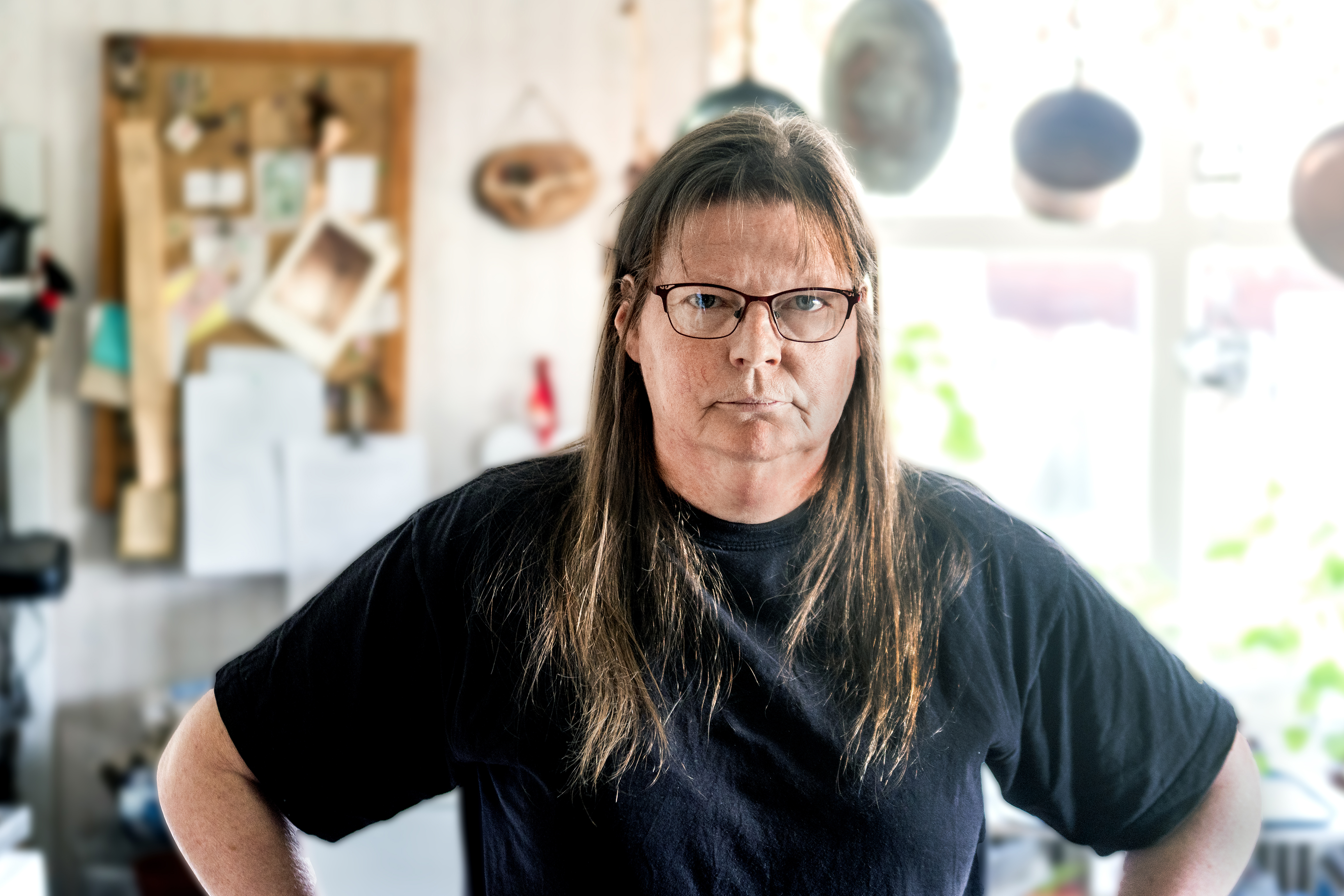Barfota Lena med långt brunt hår och glasögon står inomhus i en svart tröja med händerna vid sidorna. I bakgrunden syns ett kök med olika föremål.