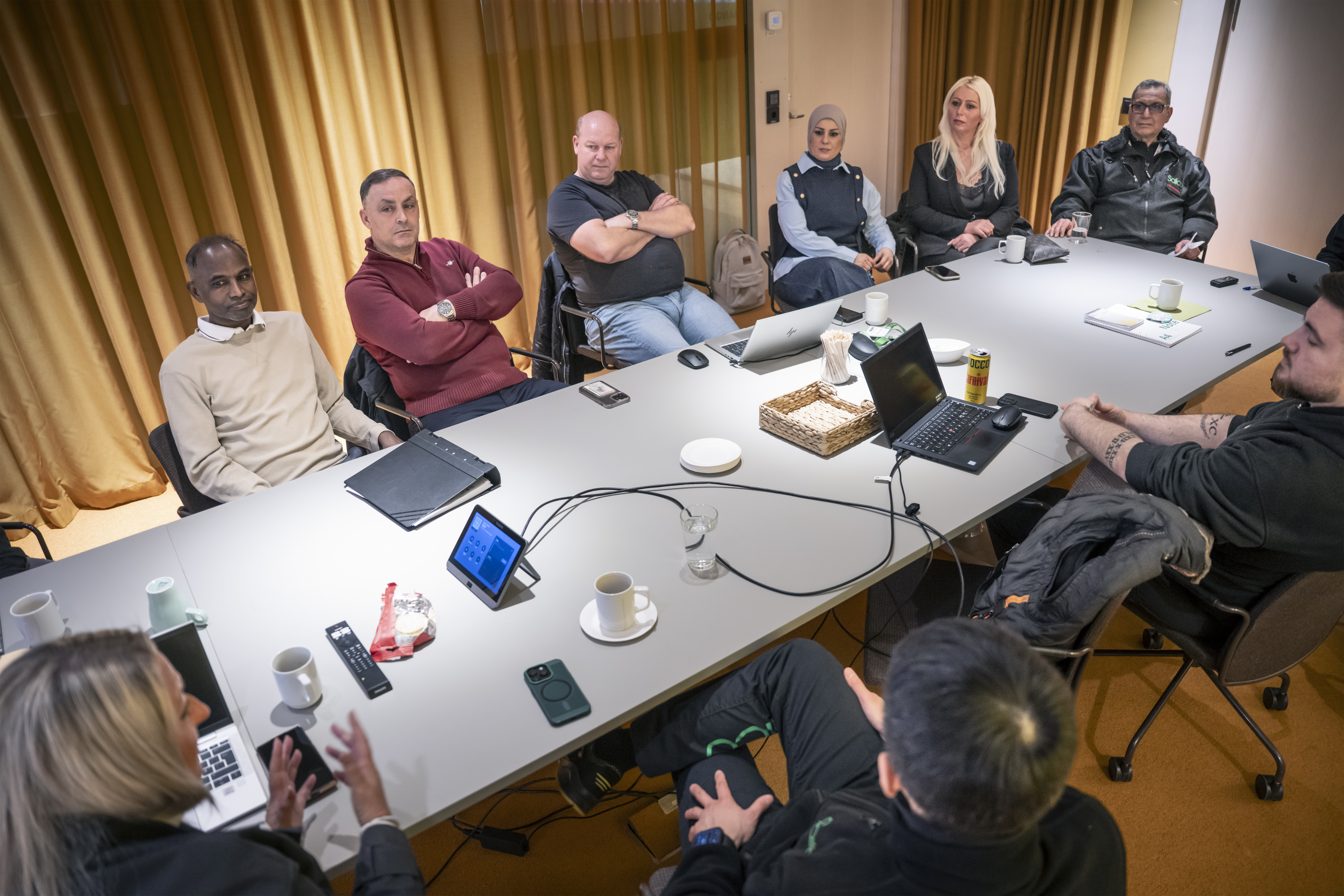 Under ett möte sitter 10 personer runt ett konferensbord med bärbara datorer, anteckningsblock och kaffekoppar.