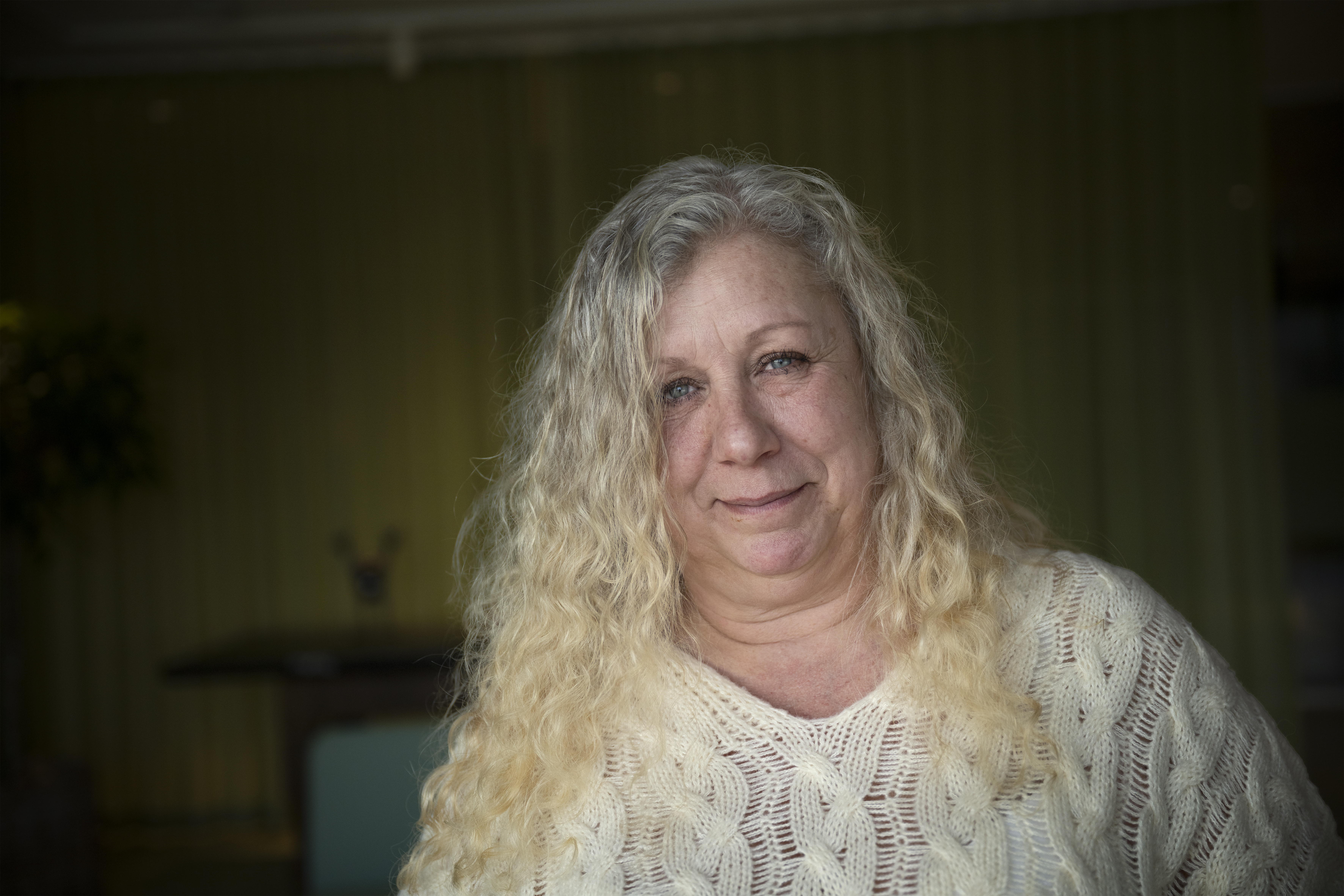 En kvinna med långt lockigt blont hår tittar in i kameran i en ljus stickad tröja och står mot en suddig grön bakgrund.