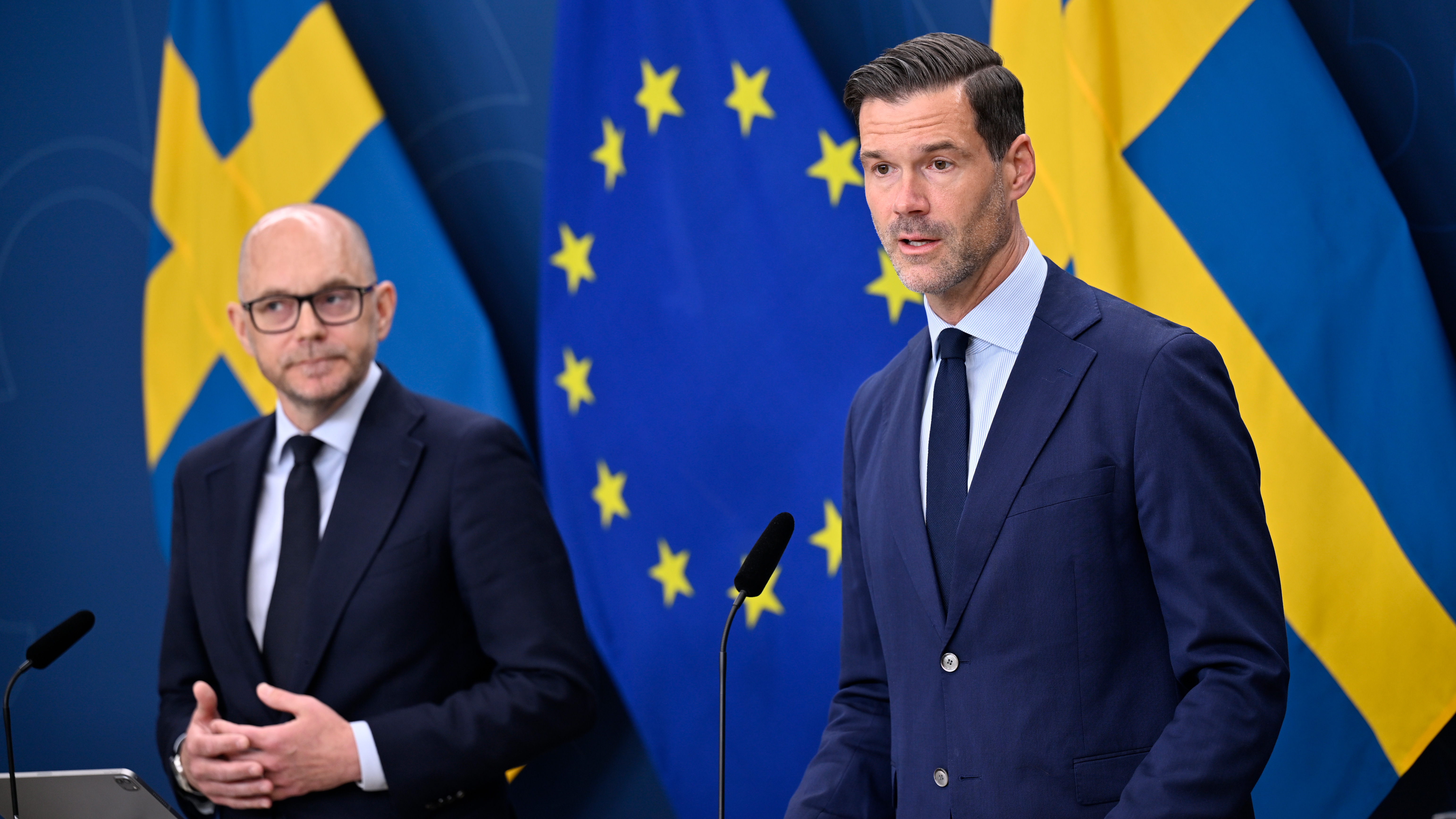 Två kostymklädda män står på ett podium framför svenska och EU-flaggan och talar på en presskonferens.
