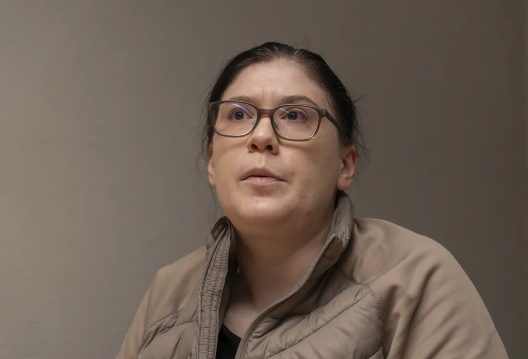 En svarthårig kvinna med glasögon är klädd i en beige jacka och tittar lätt uppåt med ett androgynt uttryck mot en enkel bakgrund.