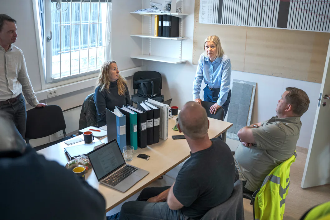 Fem personer möts runt ett bord med bärbara datorer, pärmar och kaffemuggar. En kvinna står och pratar medan de andra lyssnar.