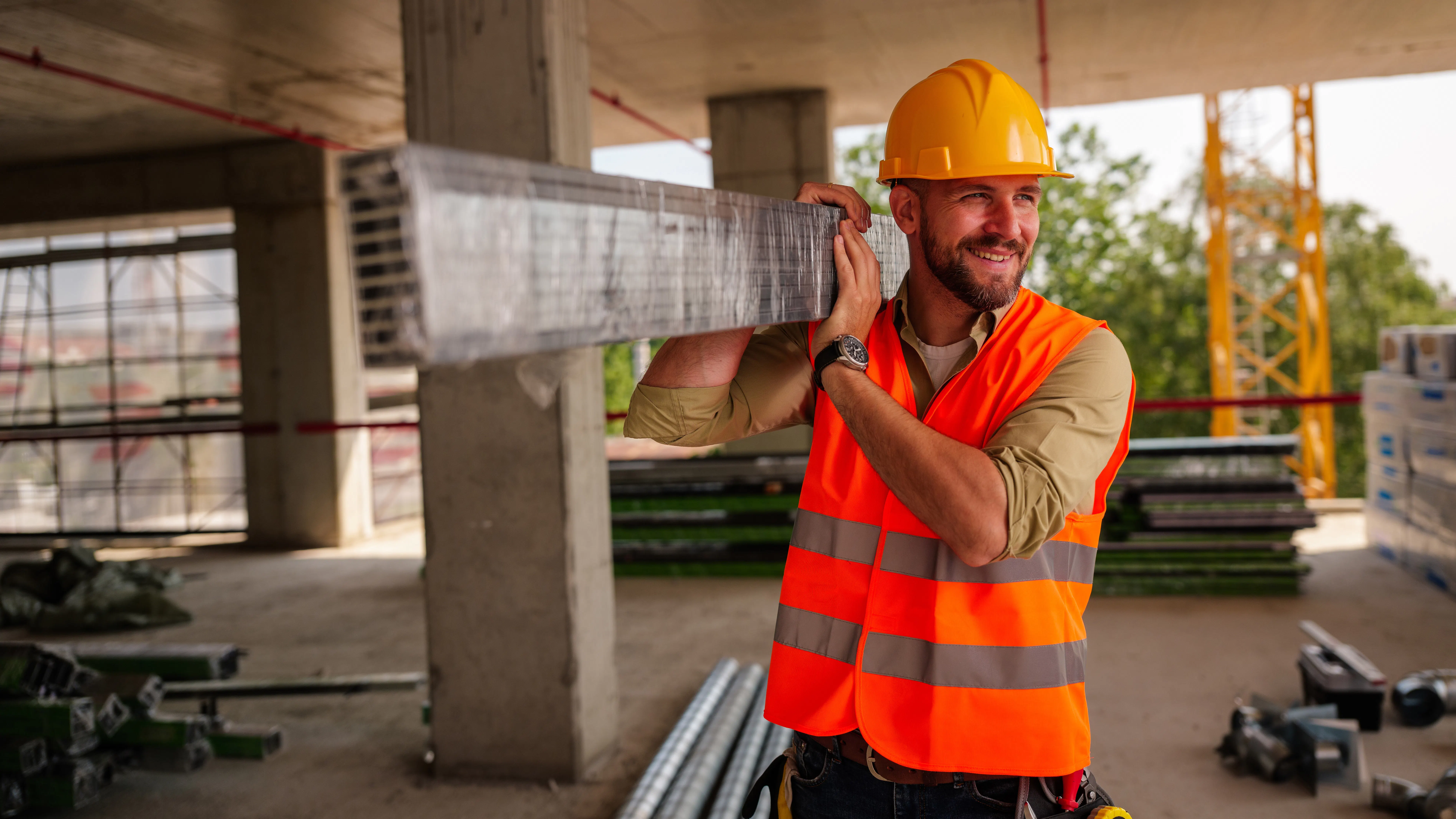 En byggarbetare i orange säkerhetsväst och gul hjälm bär en metallbalk på axeln inne i en ofärdig byggnad.