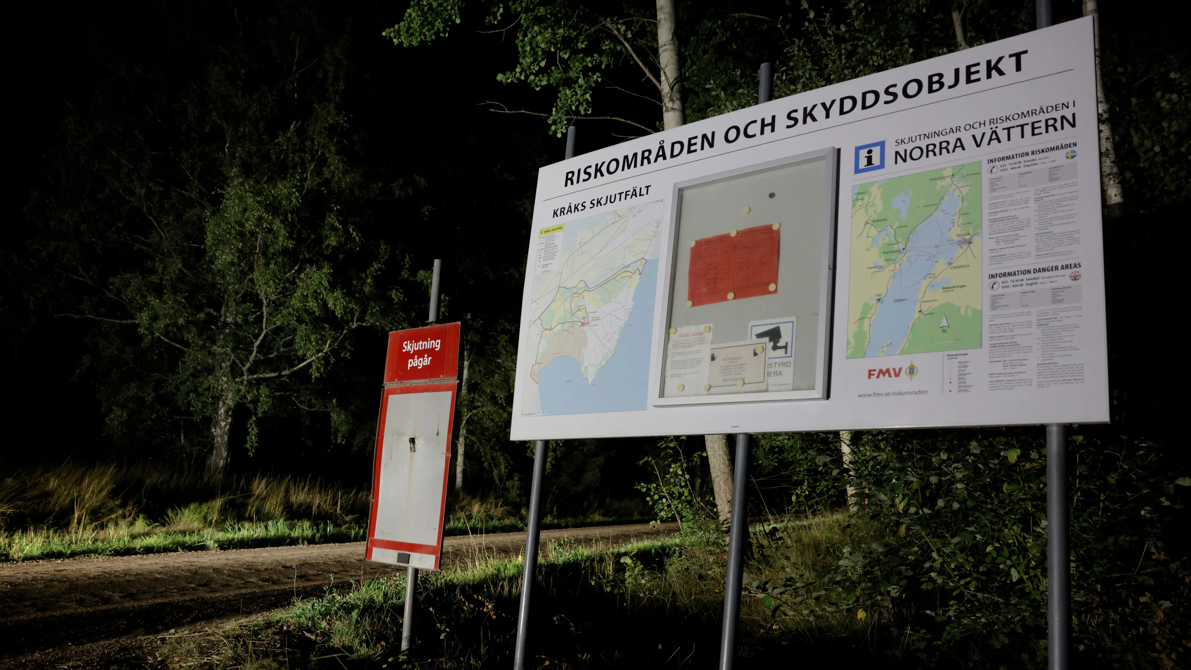 En informationsskylt med kartor och varningar står nära ett skogsområde på natten, upplyst av konstgjort ljus.