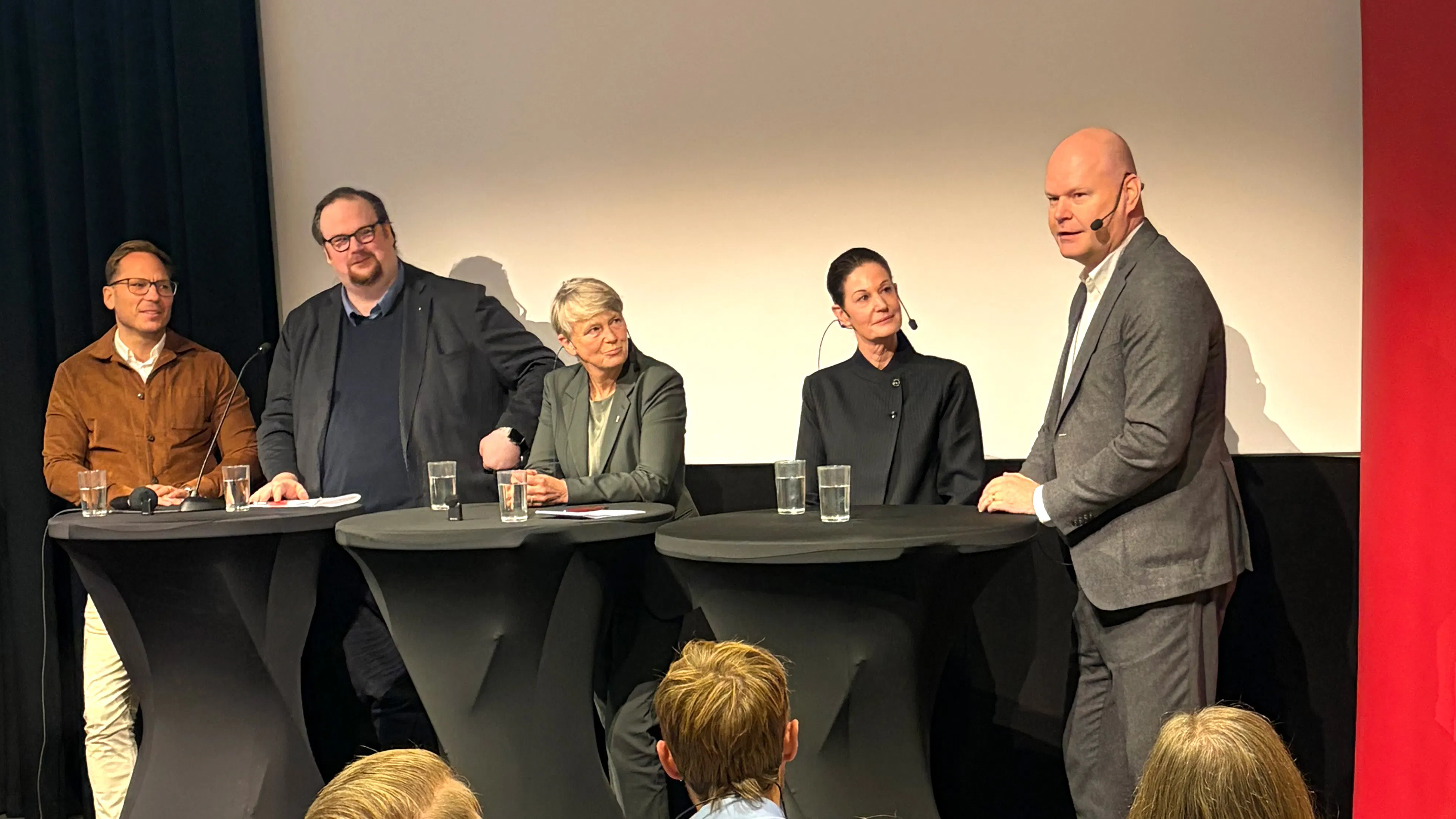 Fyra personer sitter vid ett bord på en scen medan en man står och talar till publiken; alla har mikrofoner.