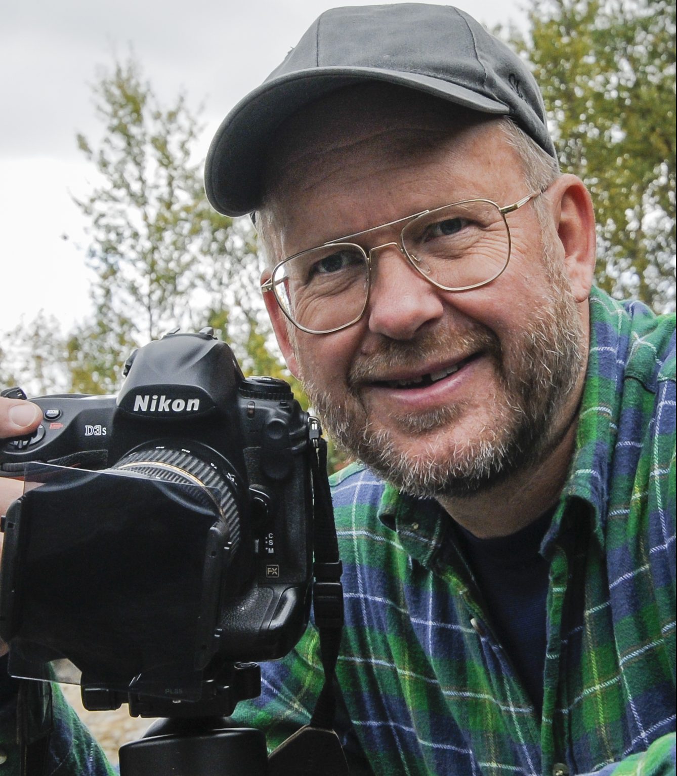 Rolf Segerstedt i keps och rutig skjorta bakom sin kamera på en klippa intill vatten.