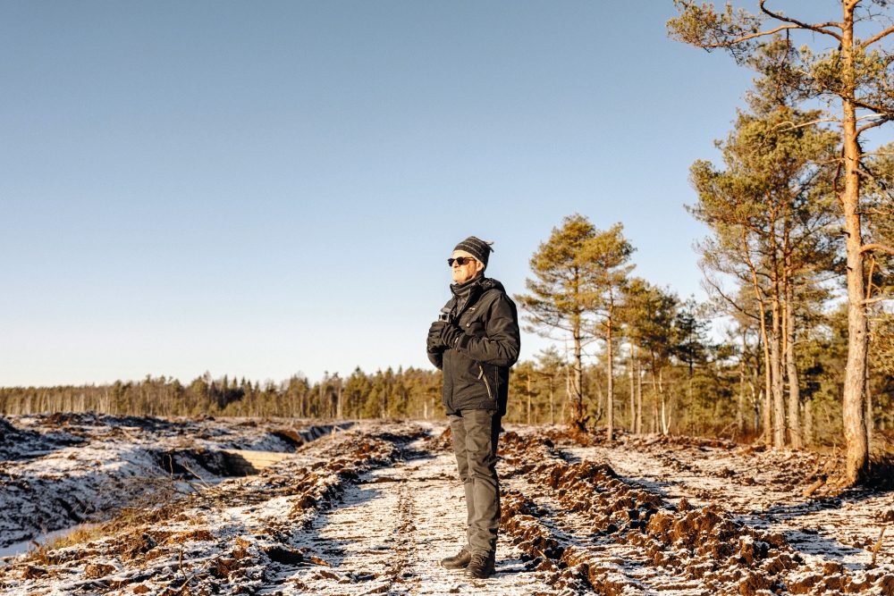 Man med svart mössa och kikar på snötäckt mosse.
