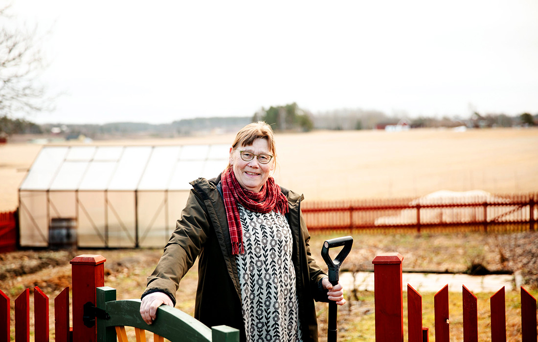 Kvinna som håller i en spade och står vid ett staket med en åker i bakgrunden.