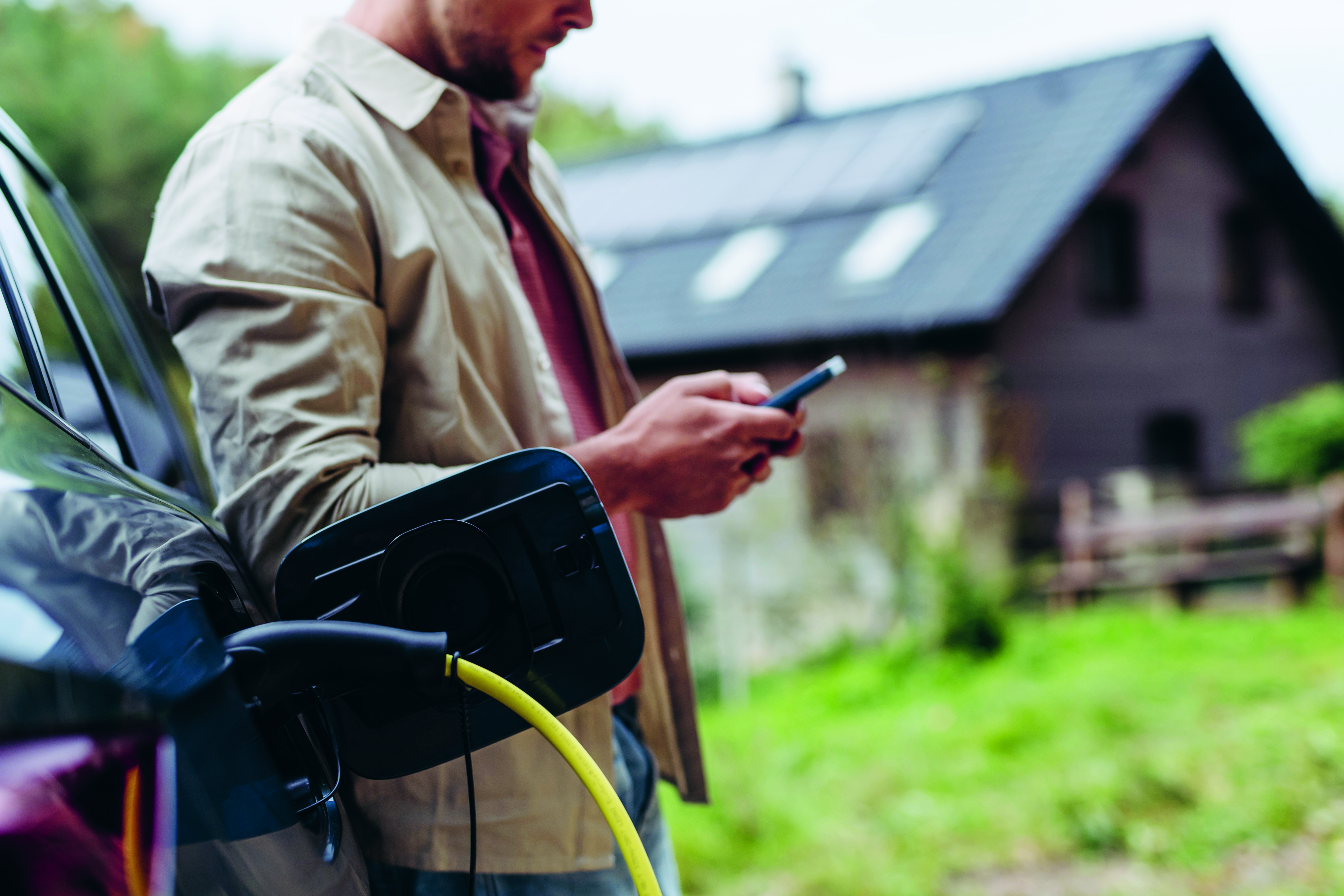 Man leaning against electric car holding smartphone in hand. Electric vehicle with charger in charging port.