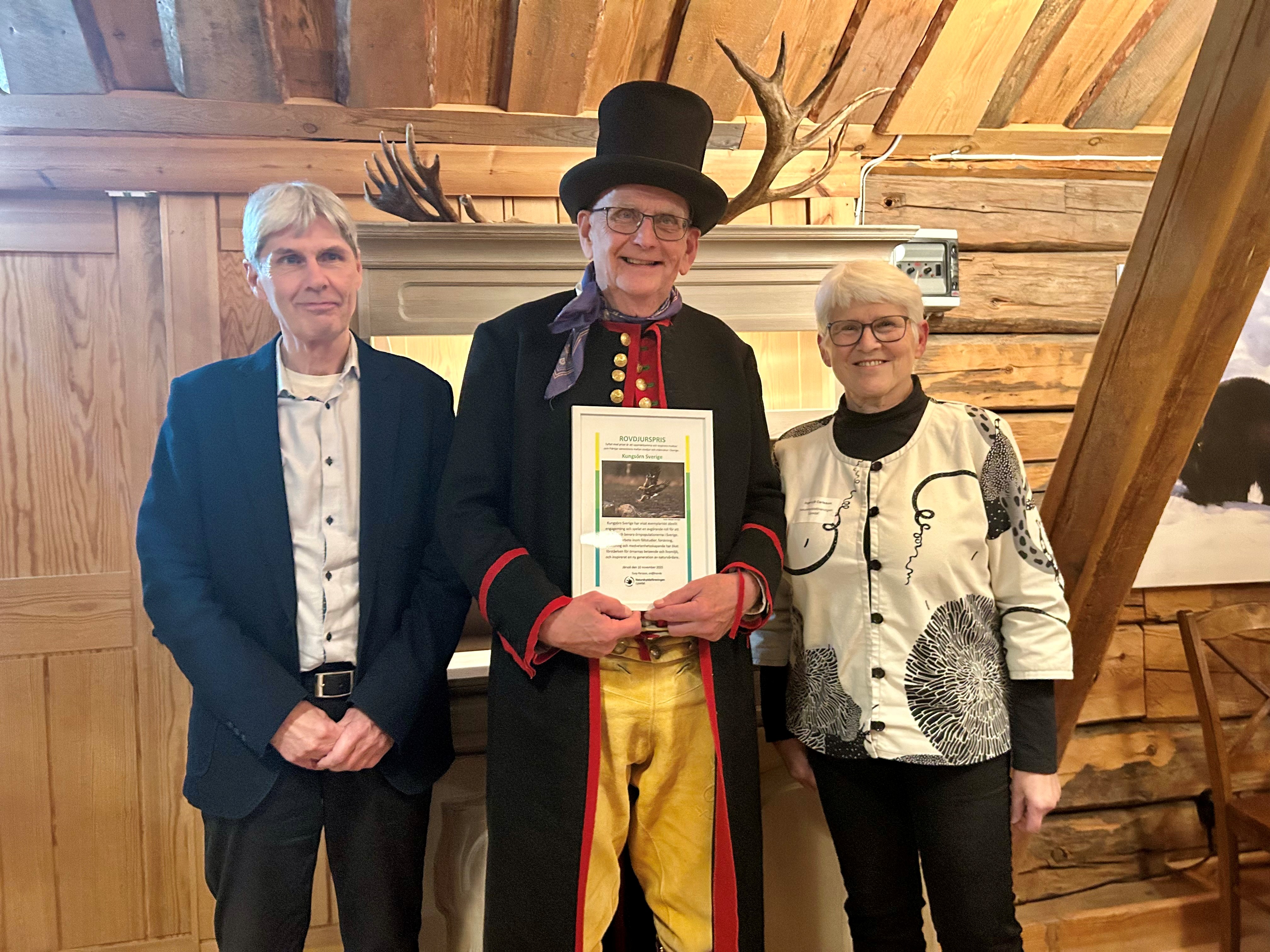 Man i folkdräkt med diplom, tillsammans med man och kvinna.