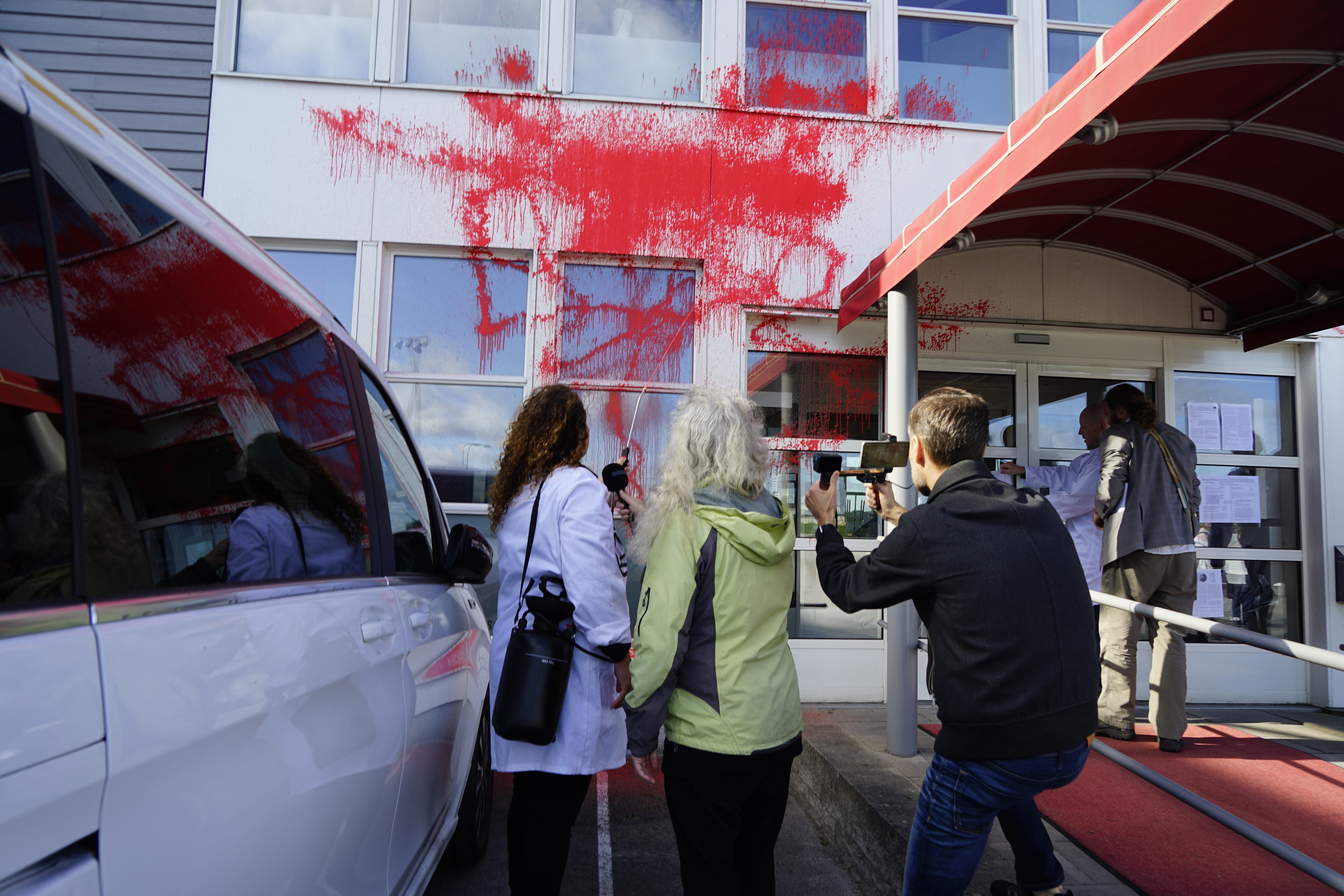 Flera personer står utanför en byggnadsentré med rött färgstänk på vägg och fönster, medan en person filmar med mobil.