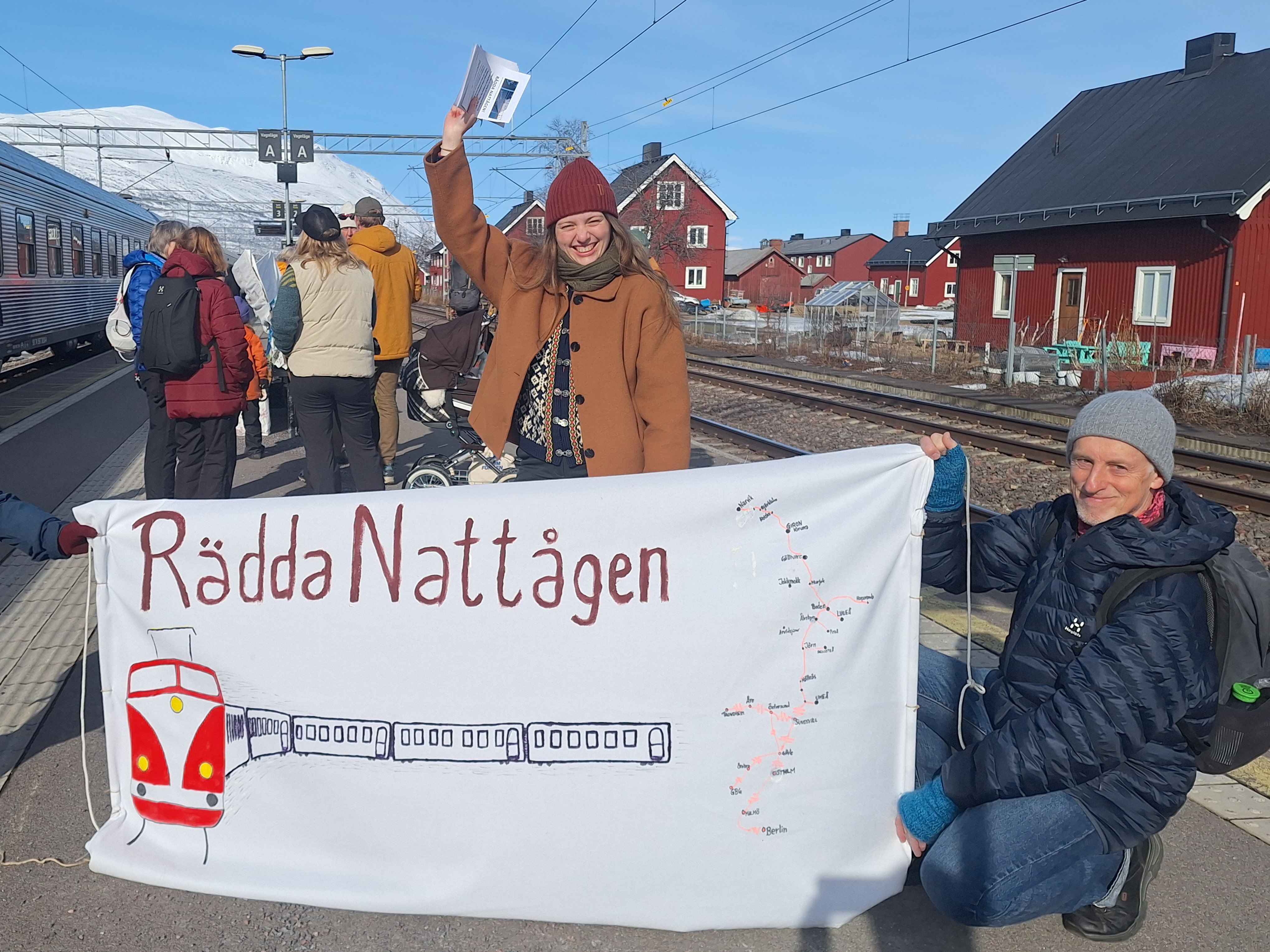 En kvinna och en man syns intill en banderoll där det står rädda nattågen på en tågperrong.