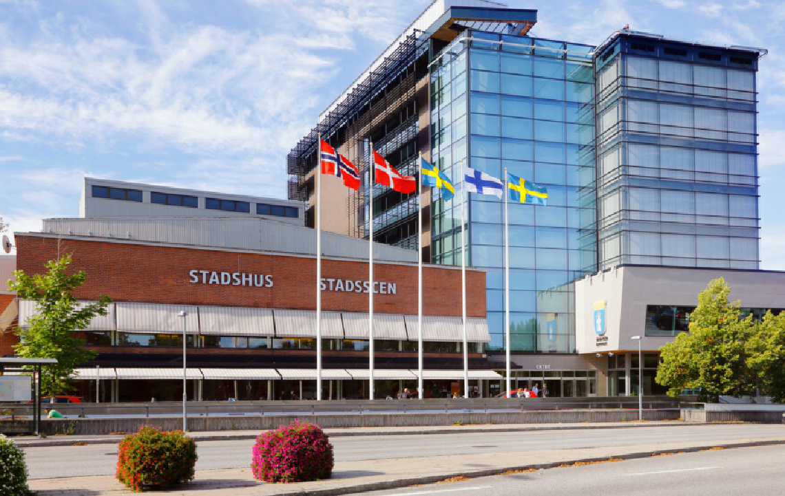 Två högre byggnader av glas och en lägre byggnad av tegel. Nordens flaggor i förgrunden.