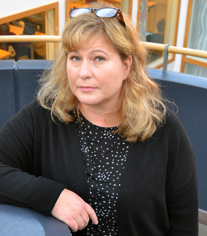 Kvinna med ljust axellångt hår och lugg, glasögon uppe på huvudet, prickig svartvit blus och svart kofta.