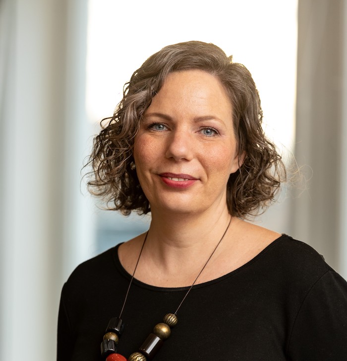 Kvinna med ljusbrunt lockigt hår i sidbena, svart tröja och ett halsband med stora trädetaljer.