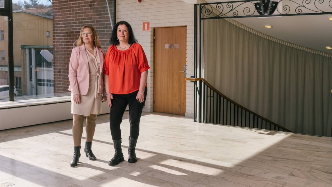 Två kvinnor står inomhus i en tom sal i solljuset som lyser in genom fönstret.