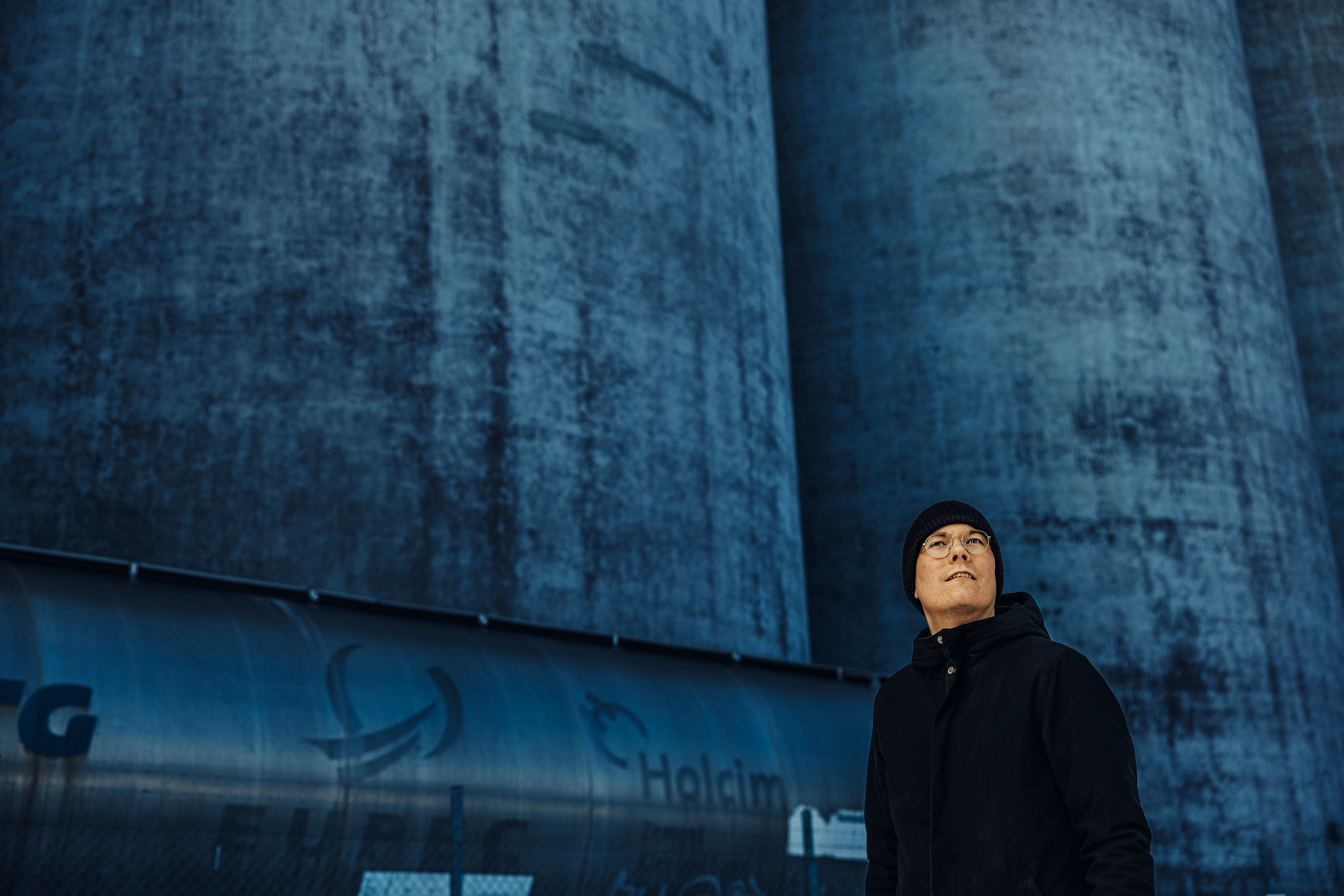 En person i svart rock och mössa står framför industriella betongkonstruktioner med en tank märkt "Holcim" i bakgrunden.