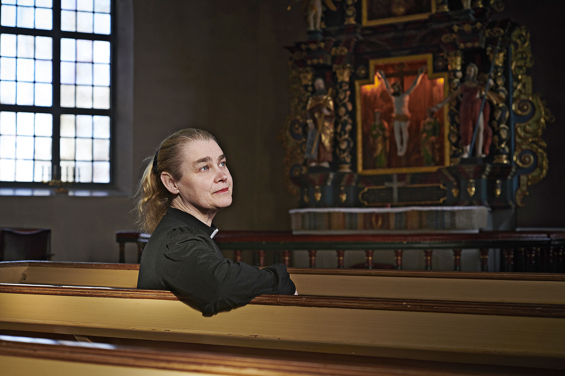 En person i prästkläder sitter i en kyrkbänk och ser eftertänksam ut. Ljus kommer in genom ett fönster. Ett altare syns bakgrunden.