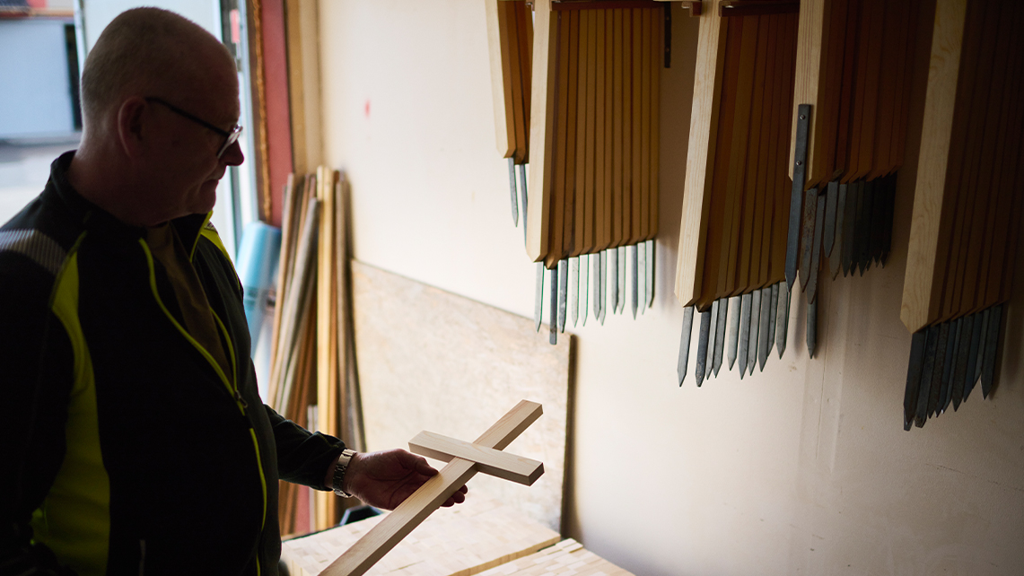 En person håller ett träkors och står nära en vägg där flera träpinnar hänger i en verkstad.